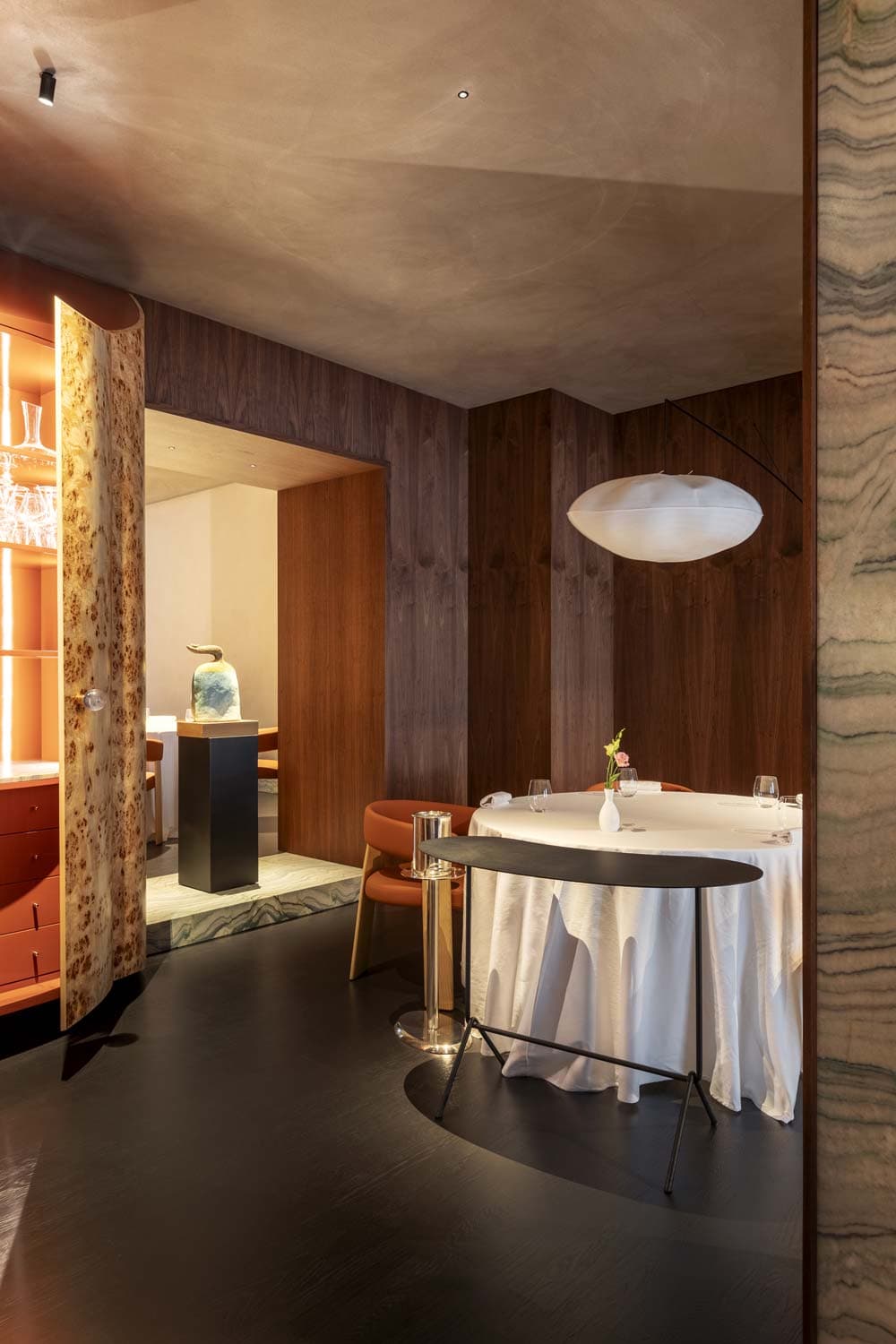 The burr-wood drinks cabinet at La Madia opened between two dining rooms — turned veneer doors in figured walnut, a glass-shelved interior of stemware, the second dining room glimpsed through the opening with its onyx-marble plinth and a hand-painted Sicilian ceramic.