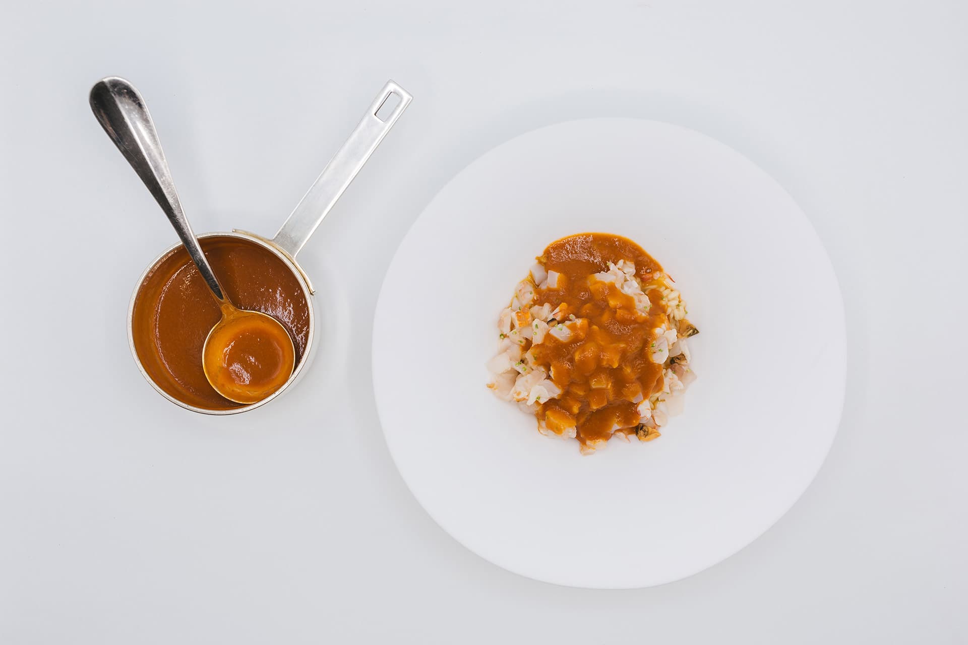 An overhead plate of Pino Cuttaia's seafood ragù — small white discs of squid and crustacean dressed with a glossy ochre tomato sugo, served beside a steel pot of the same sauce on a long thin handle.