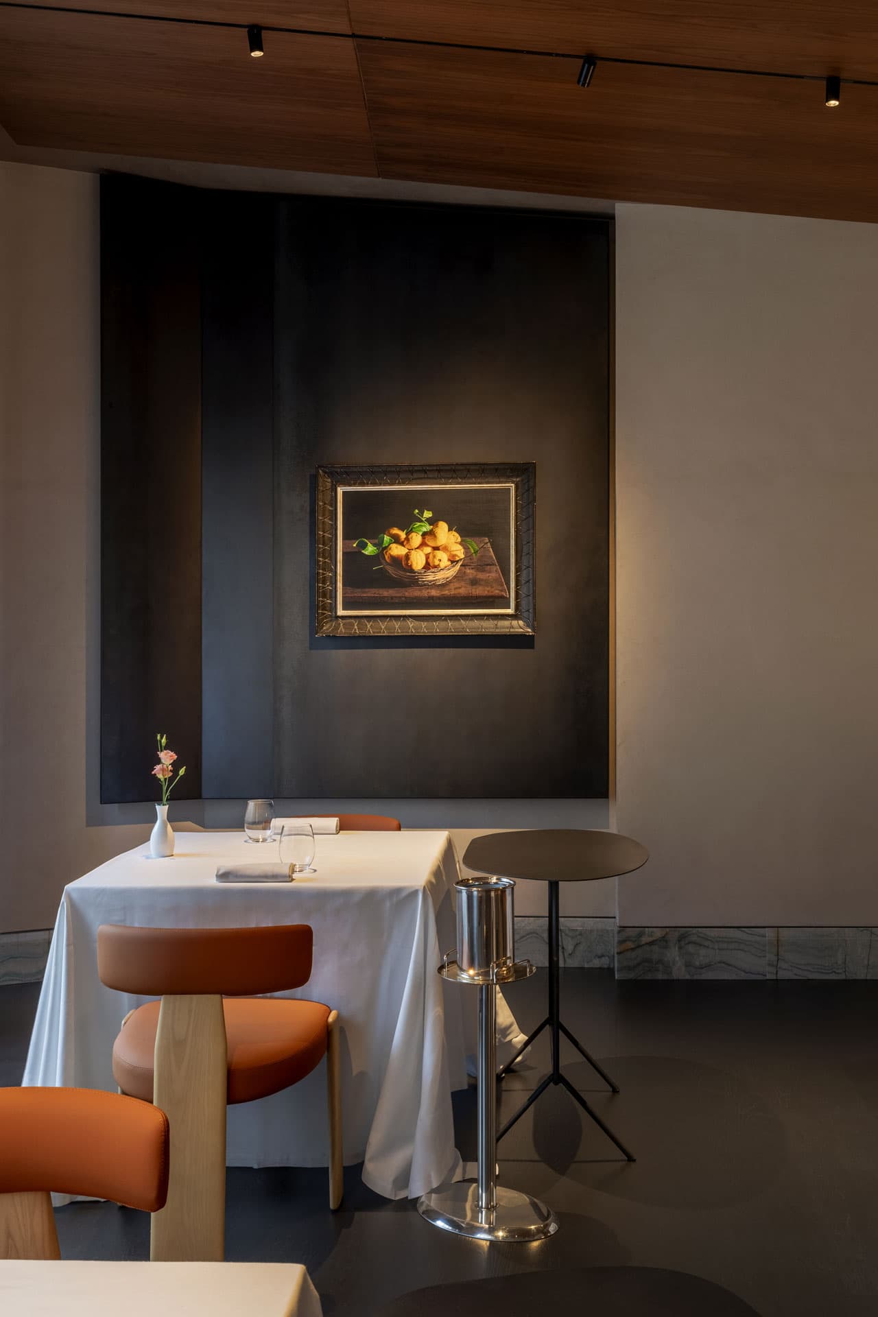 A corner of La Madia's dining room — a single white-clothed table set with a pink lisianthus and a crystal stem, terracotta chairs in pale ash, and an oil painting of a basket of golden Sicilian lemons hung over a panel of burnished black iron.