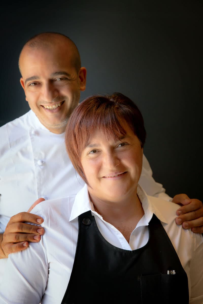 Pino Cuttaia and Loredana Cipriano photographed in profile against a black ground — the chef in chef's whites with his hands on his wife's shoulders, Loredana in a black apron over a white shirt, both smiling.
