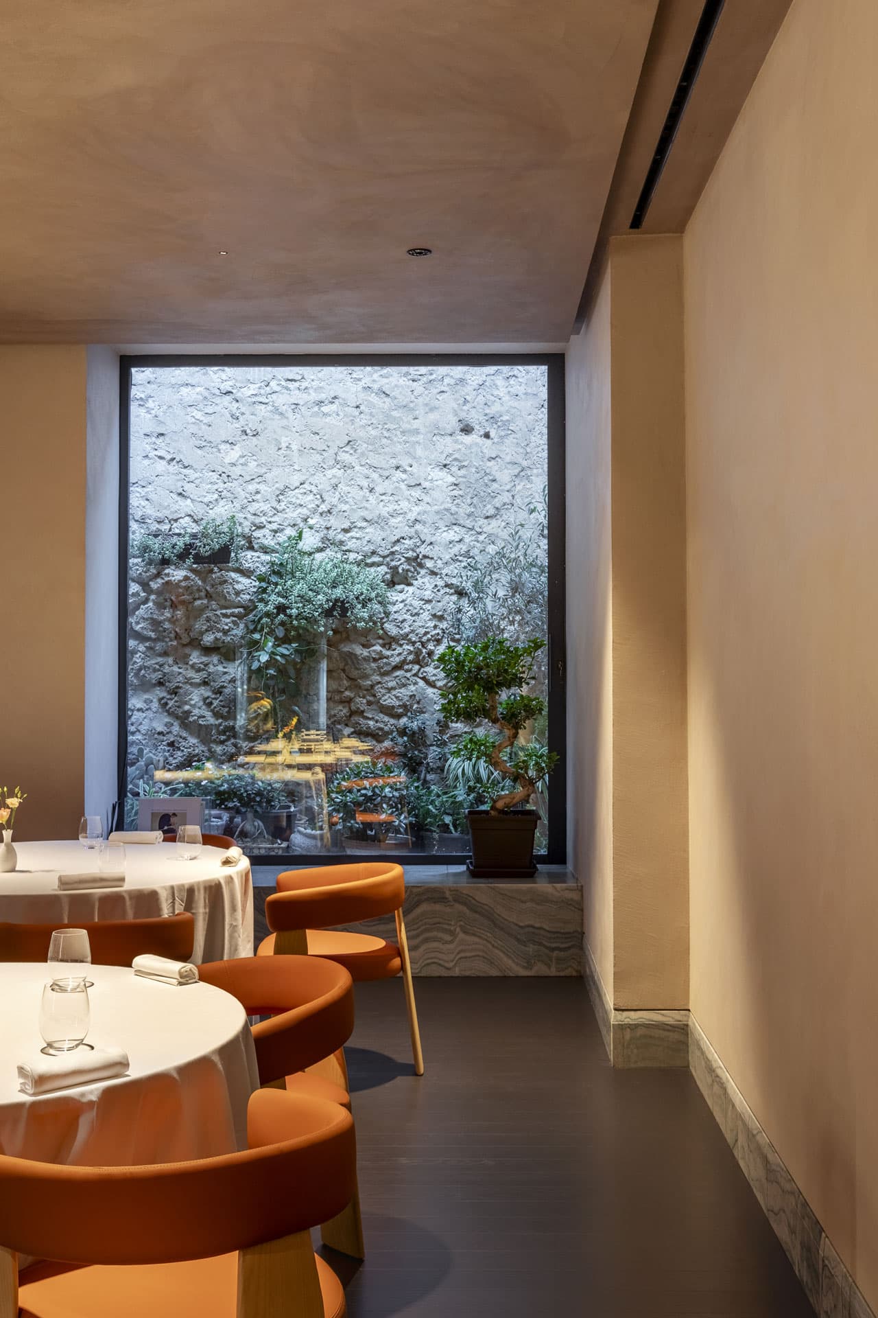 An intimate corner of La Madia — terracotta chairs around a small round table, a tall window opening onto an inner courtyard of bare ochre stone with a single small bonsai and a clutch of Sicilian succulents.