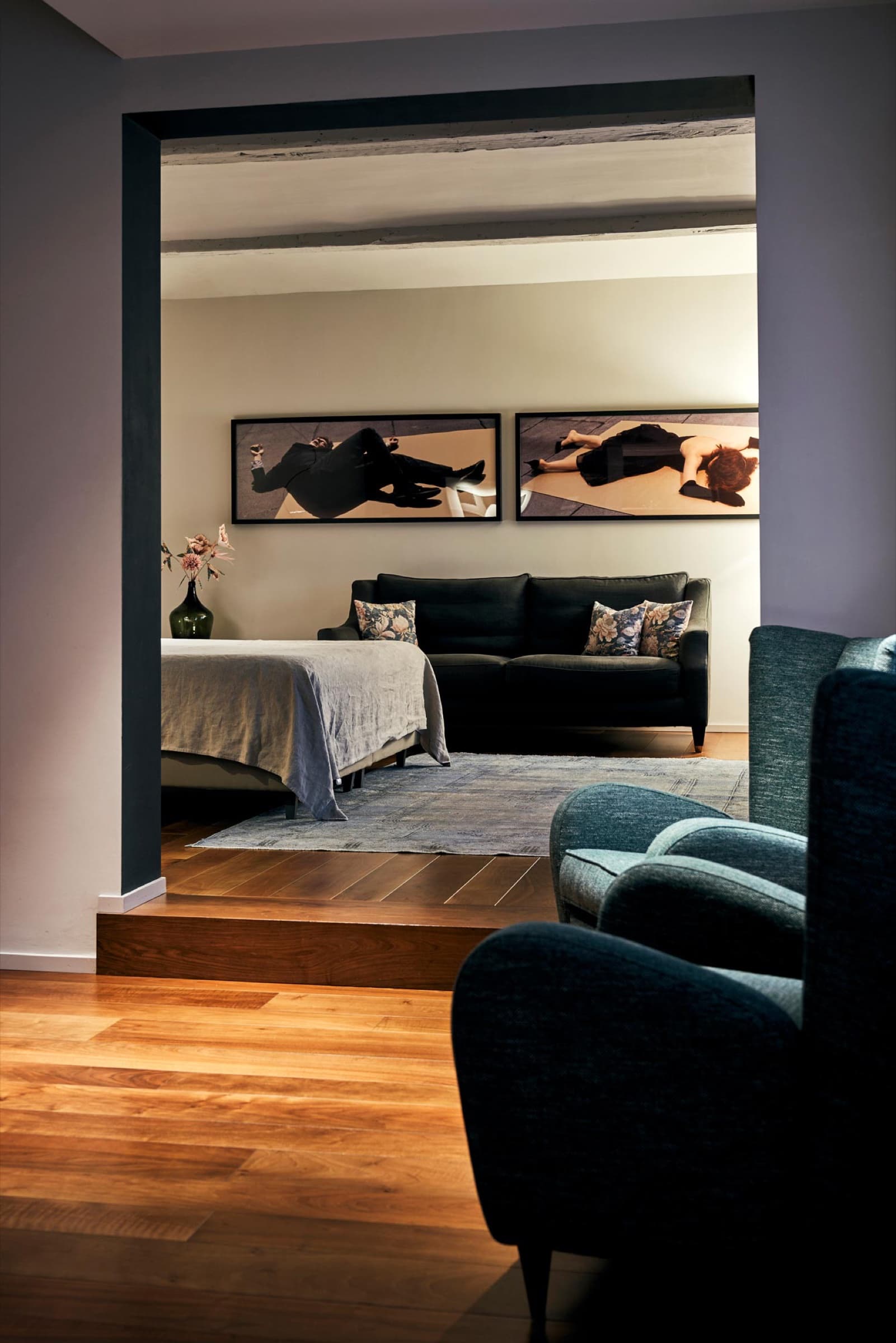 A guest suite seen through an open doorway — two large dark photographic prints above a charcoal sofa, a linen-dressed bed, a teal armchair in the foreground.