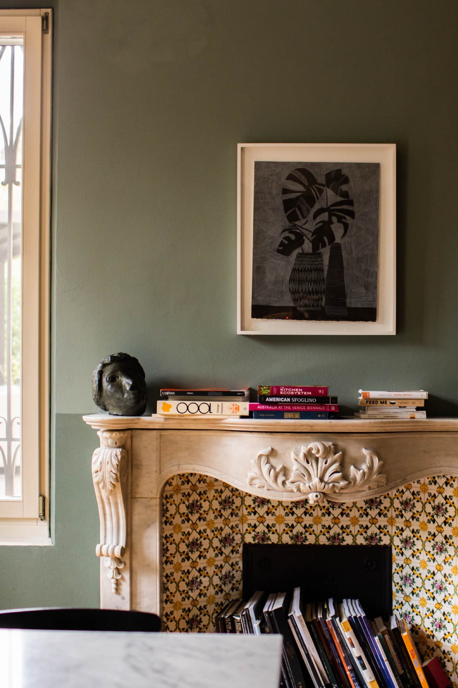 A sage-green sitting room corner — an antique marble mantel with a yellow-tiled fireback, a contemporary print of a vase above, design and art books stacked on the shelf below.