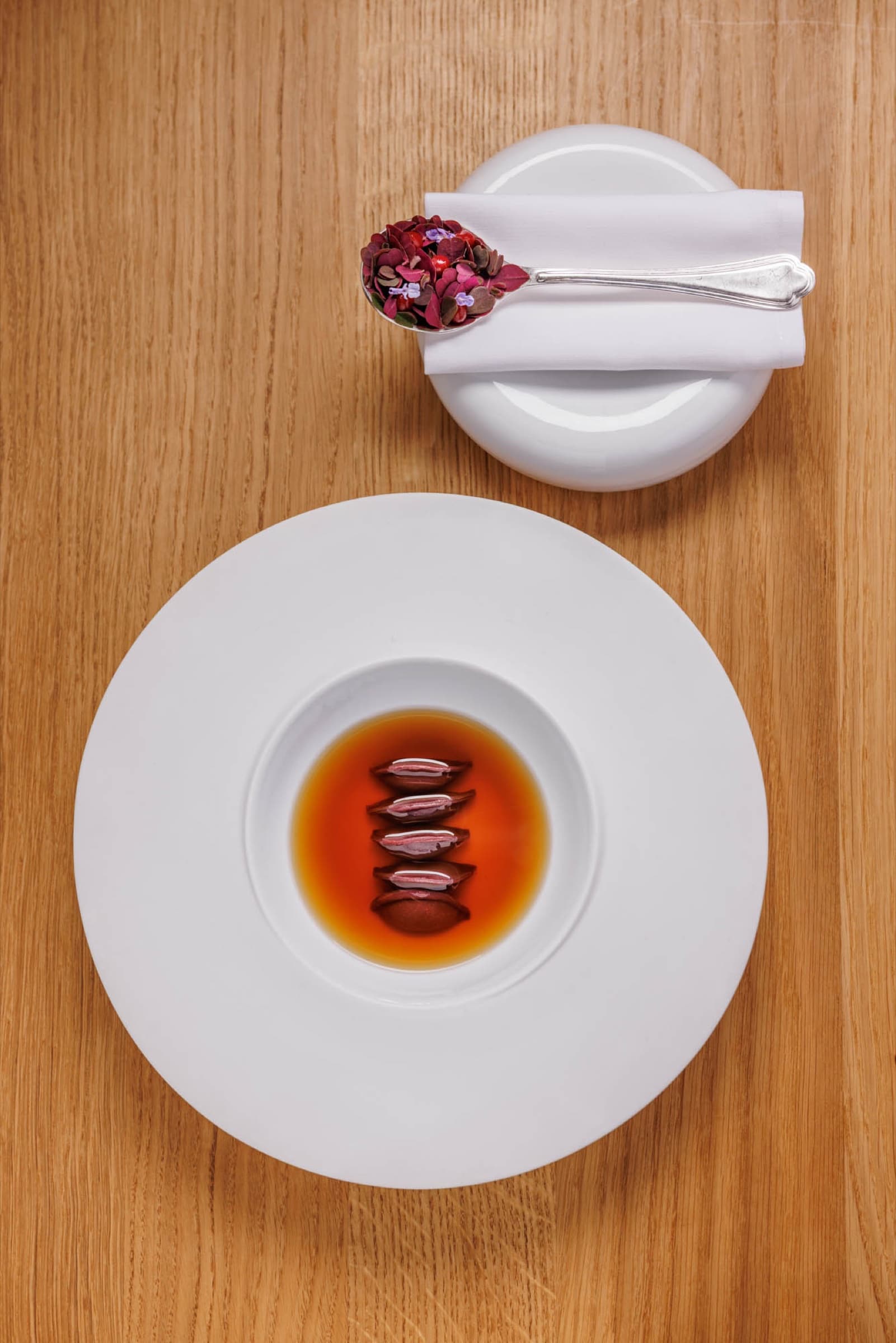 An overhead plate at Francescana at Maria Luigia — five small ravioli in a deep amber broth on a white porcelain bowl, a dish of red flower petals beside.