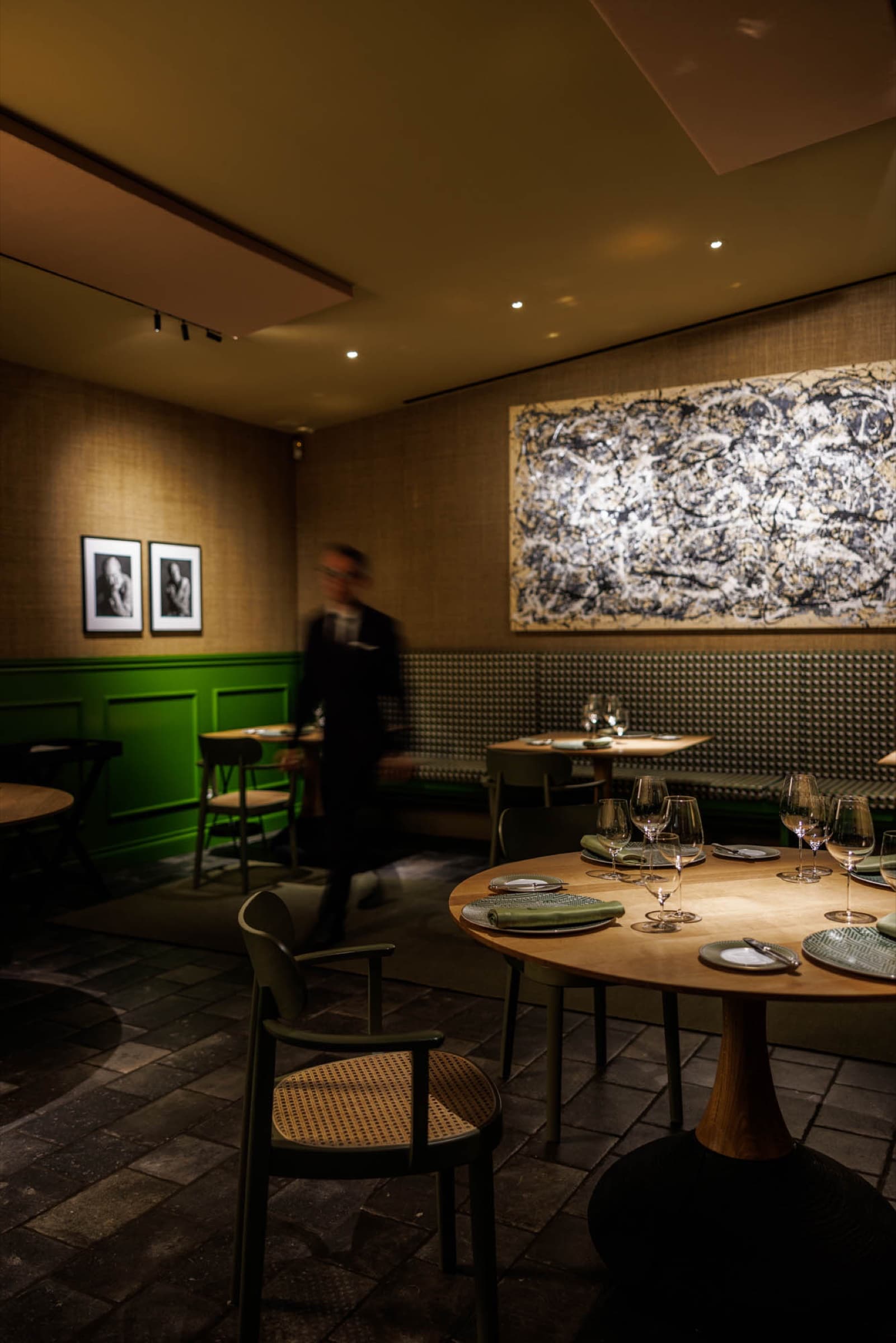 The Al Gatto Verde dining room — green panelled wainscot below natural-fibre walls, a large drip-painted canvas above a banquette, an oak round table set with green linen napkins and stemware.