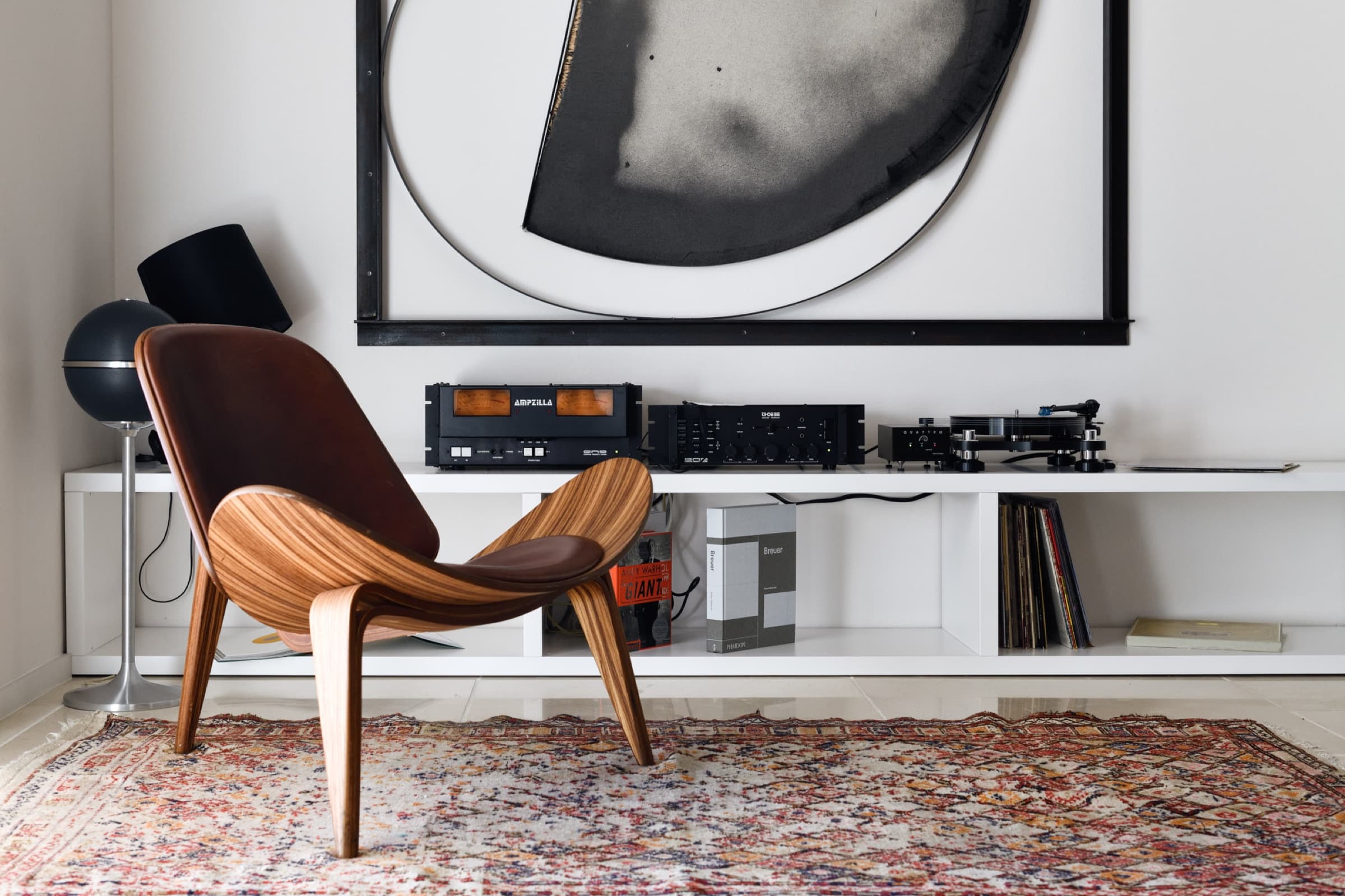 A pale interior with a Wegner Shell chair on a faded oriental rug, a vinyl turntable and amplifiers on a low white console, a large round black-and-white abstract canvas above.