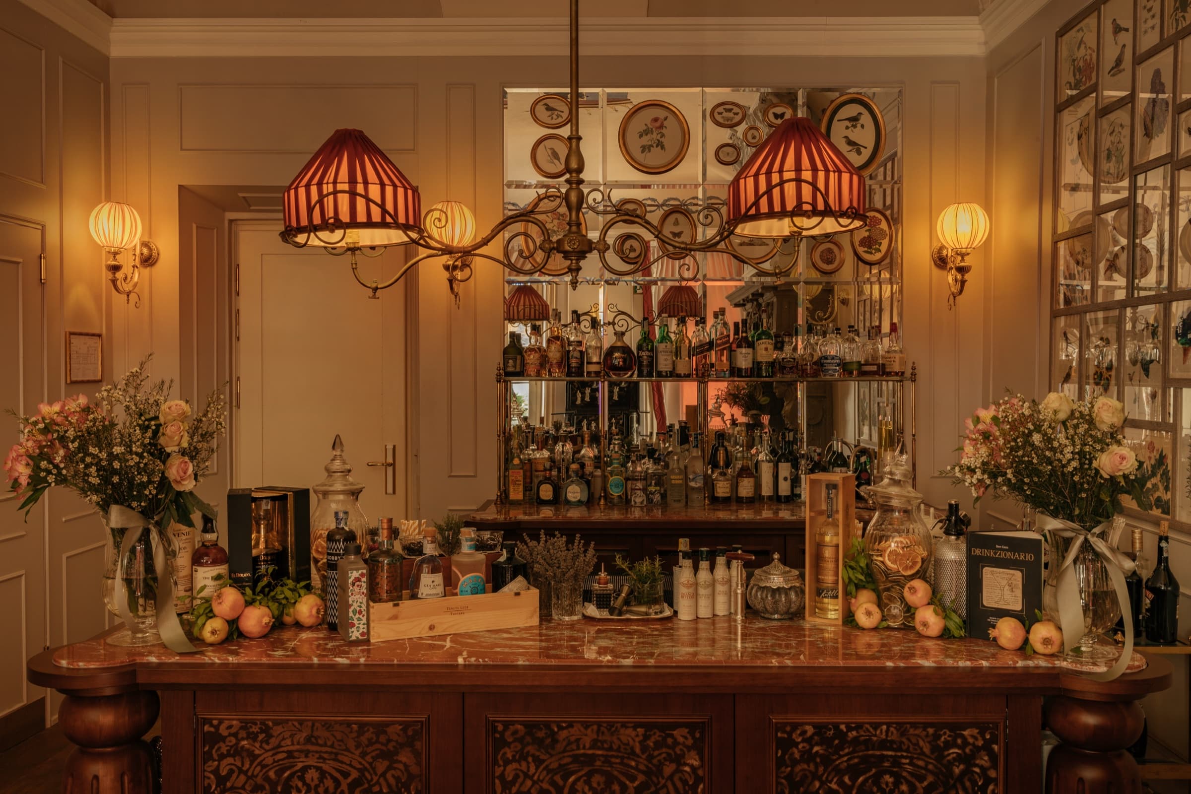 The bar at Passalacqua — wood panelling, a mirrored back, Fortuny pleated lampshades, a marble counter set with bottles, glassware and pale roses.