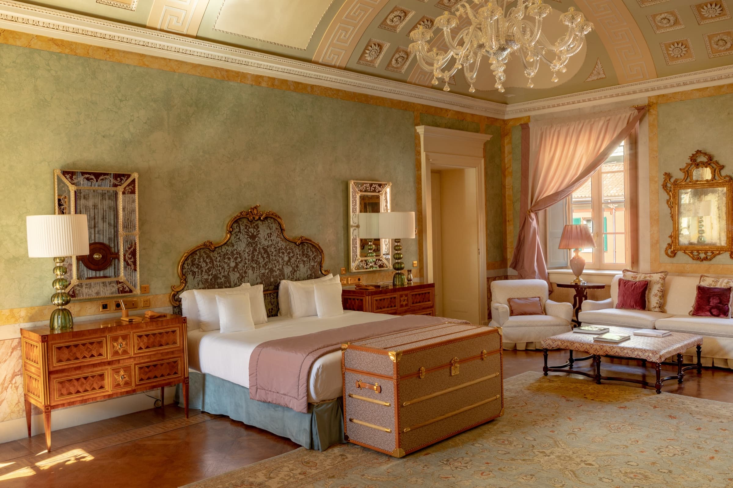 A guest suite in the Villa — sage-green walls, a gilded baroque headboard, a brass-bound trunk at the foot of the bed, a Murano chandelier above.