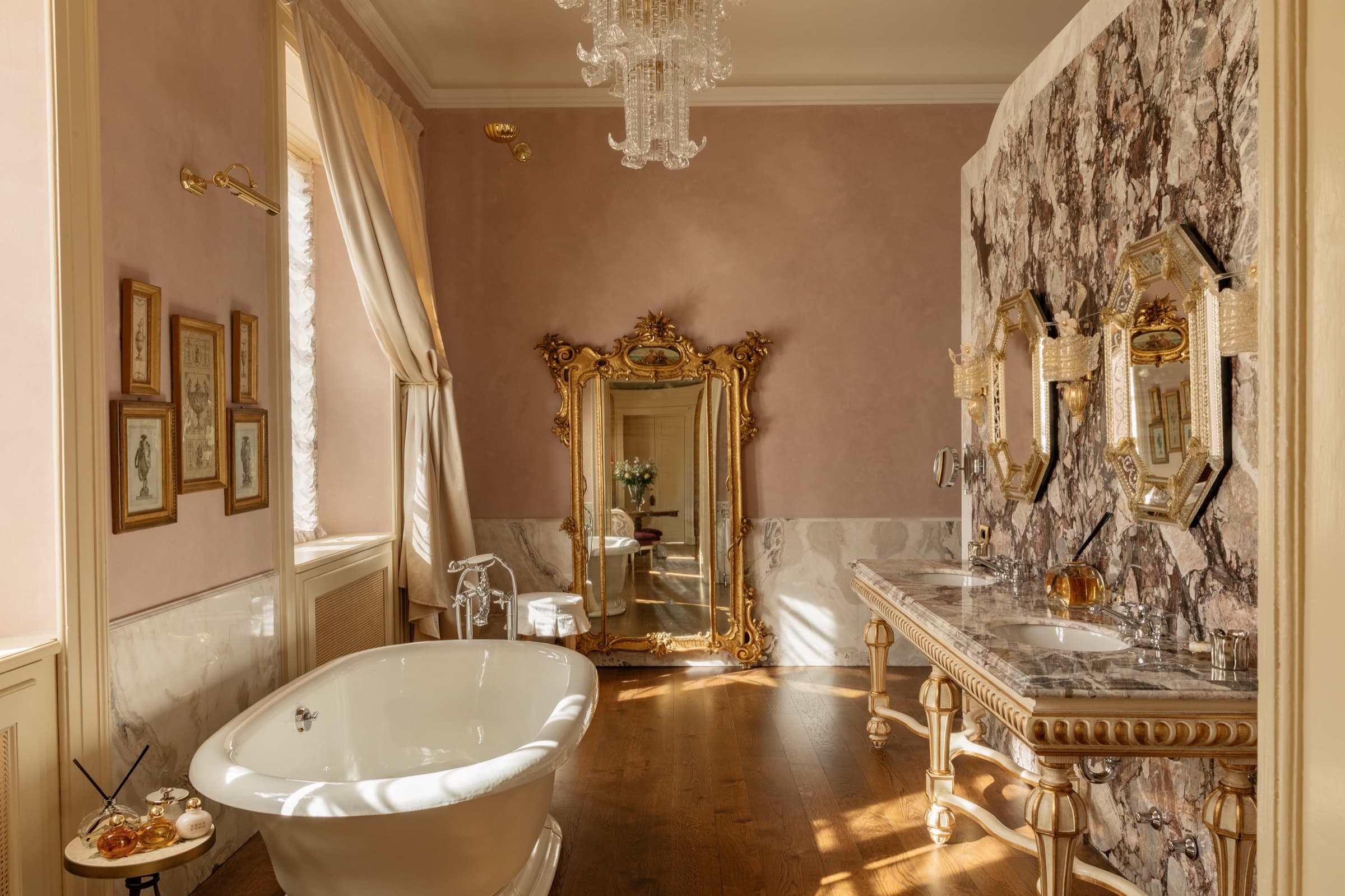A bathroom with rose-pink walls, a freestanding white bath, a double marble vanity and a gilt baroque mirror in late afternoon light.