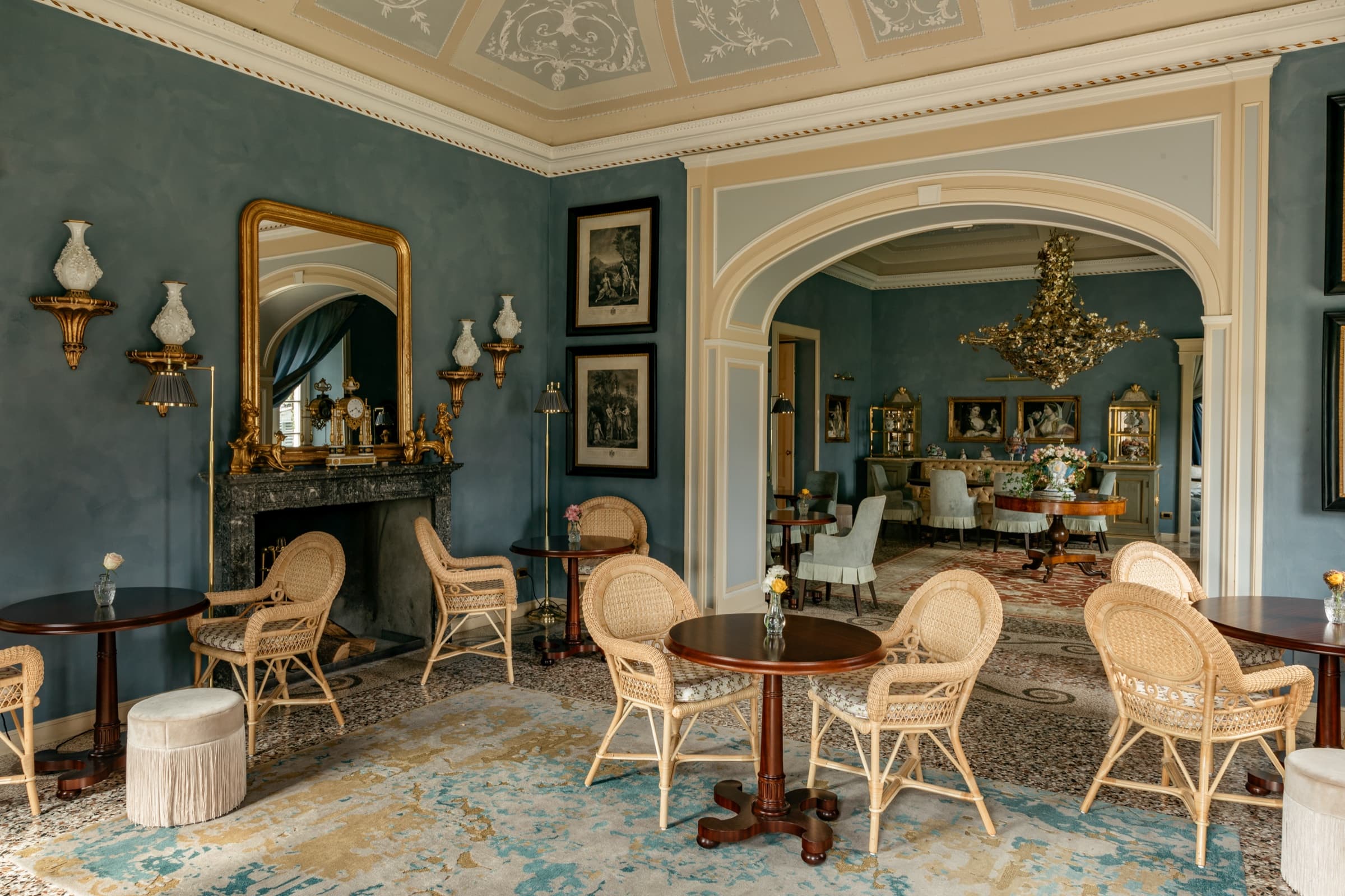 A blue salon with a marble fireplace, gilt overmantel mirror, wicker café chairs around small round tables and a worn antique rug on the terrazzo.