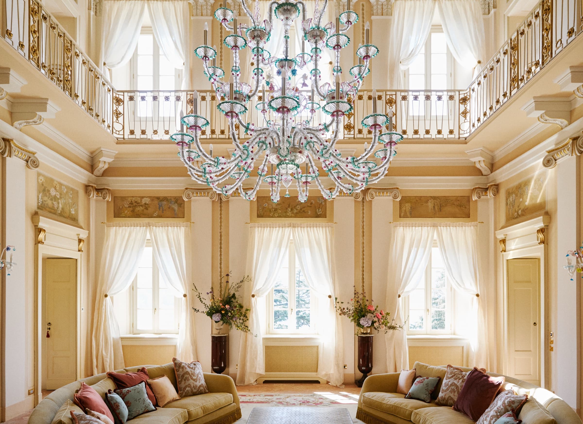 A double-height ceremonial salon with a Murano chandelier, painted upper balustrade, frescoed friezes and tall cream-curtained windows.