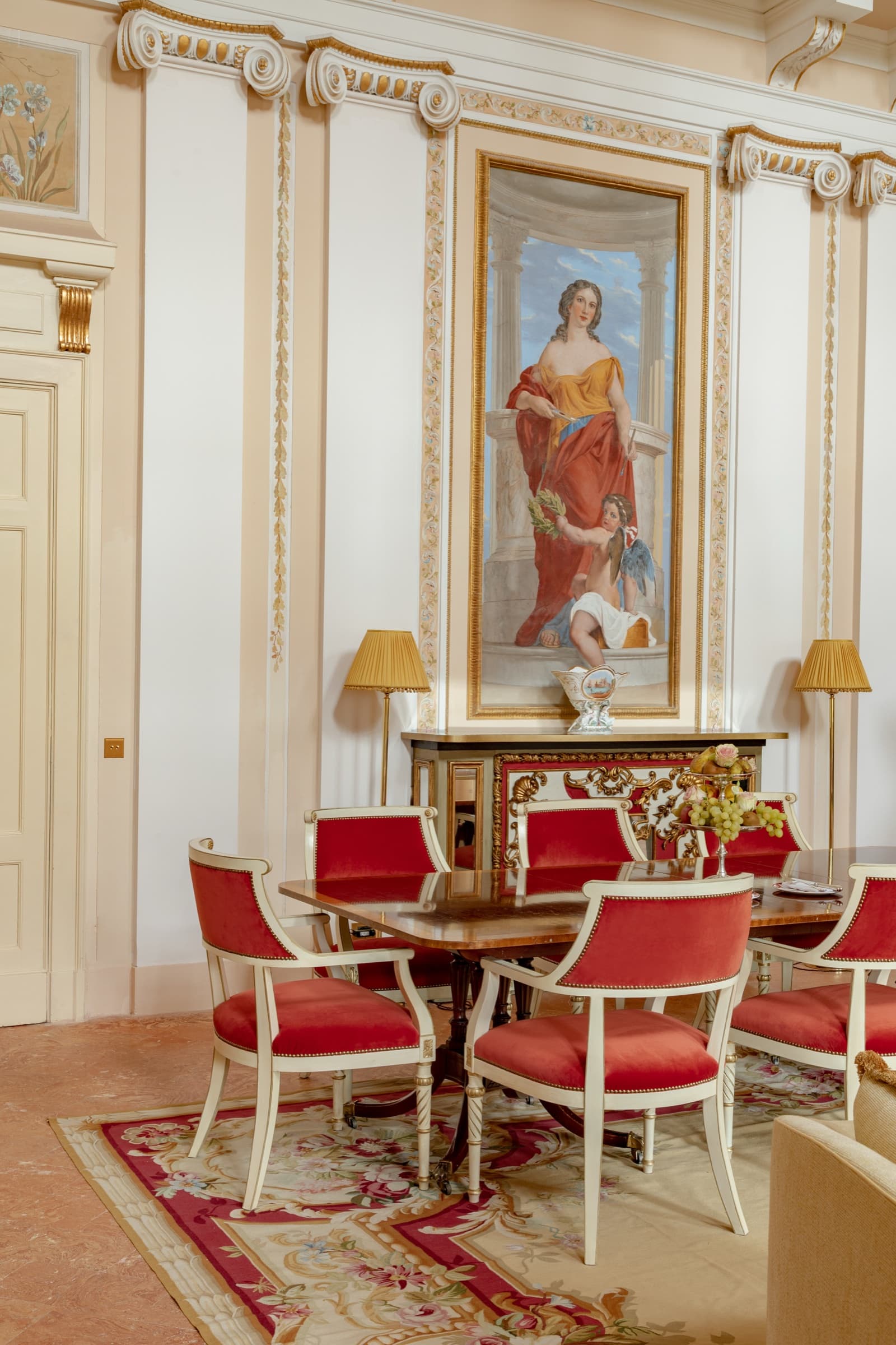 A neoclassical dining room with cream-and-gold pilastered walls, a large fresco of a draped woman, and red velvet chairs around a polished table.