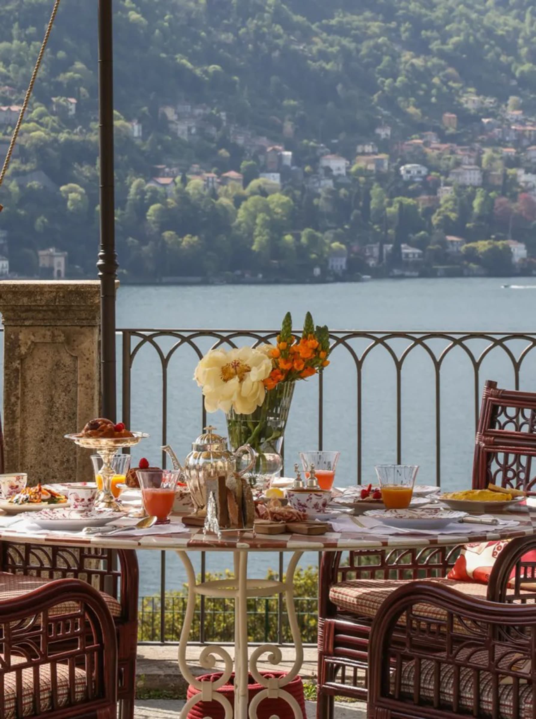 A round marble breakfast table on a wrought-iron lakeside terrace — orange tulips, a silver coffee pot, fresh pastries — Lake Como and the western shore in soft morning light.