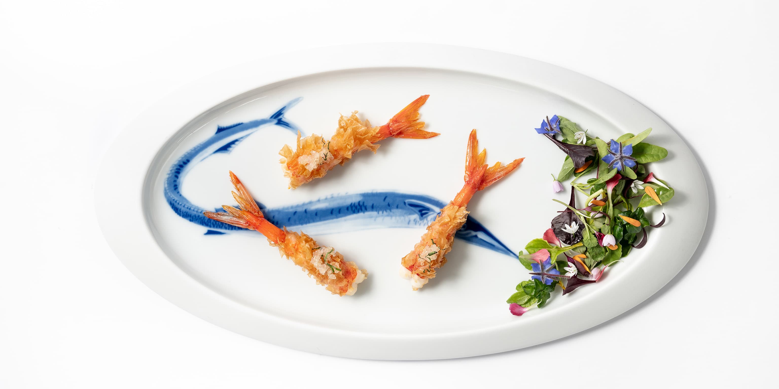 A long oval white plate at Quattro Passi — three crisped Mediterranean prawns laid in a curve over a hand-painted blue fish motif, with a small tangle of borage flowers and herb leaves at one end.