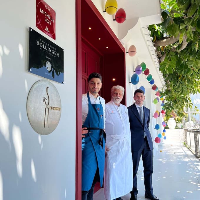 Three Mellinos at the entrance to Quattro Passi — Fabrizio in a blue apron, his father Antonio in chef's whites and Raffaele in a dark tailored jacket — standing under a Michelin 2022 plaque and a row of colour-glazed ceramic eggs hung along the white wall.