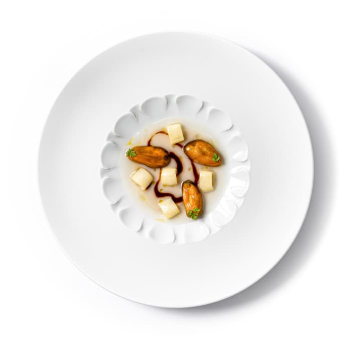 A small white scallop-edged bowl set inside a deep round plate — three plump Tyrrhenian mussels and small pale cubes in a clear shellfish broth lined with a single arc of dark vinegar, photographed from above against a bright white ground.
