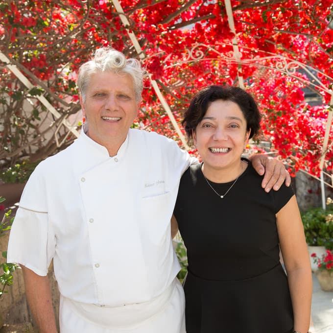 Antonio Mellino in chef's whites with his wife Rita standing under the bougainvillea arch at the entrance to Quattro Passi — both smiling, the heavy red-pink blossom blurred behind them in the midday sun.