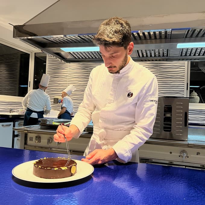 Fabrizio Mellino in chef's whites at the pass, plating a glossy chocolate tart with a small gold leaf insignia — the rest of the brigade at work in the open kitchen behind him under bright extractor light.