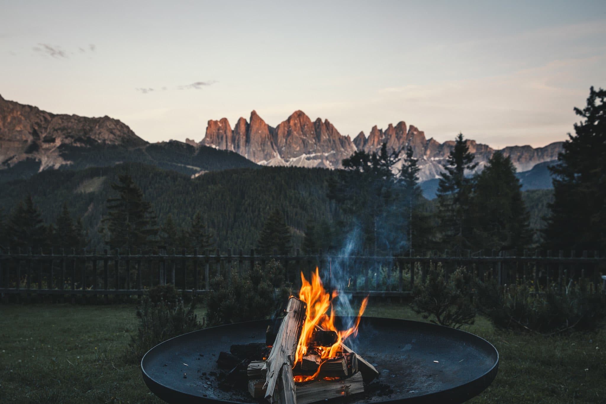 A copper firepit in the meadow at dusk, low flames against the sawtooth profile of the Geisler peaks turning rose under a clearing sky.
