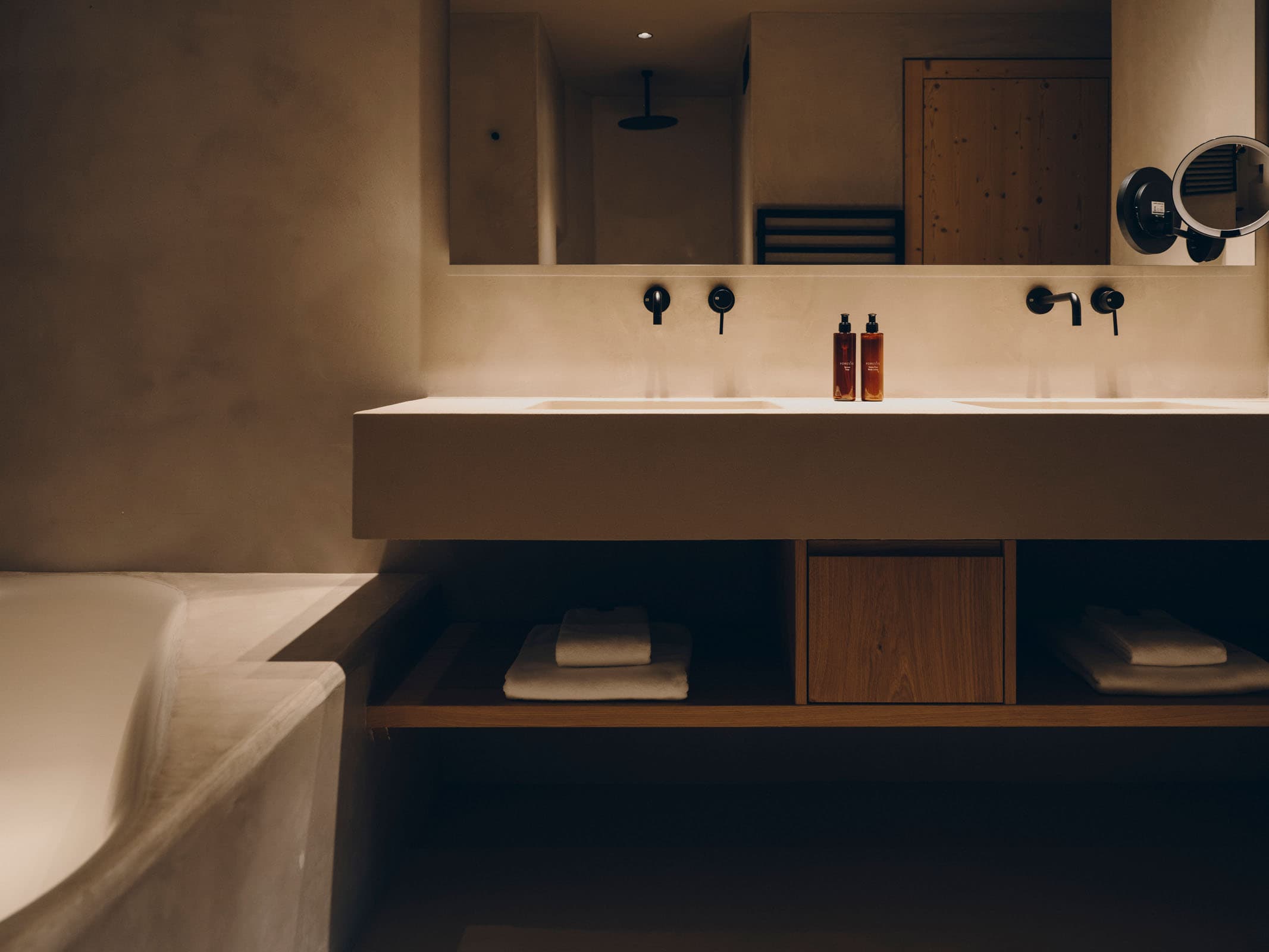 A clay-rendered bathroom in warm sand tones, stone-pine cabinetry under a long basin and a soft-edged tub at the side of the frame.