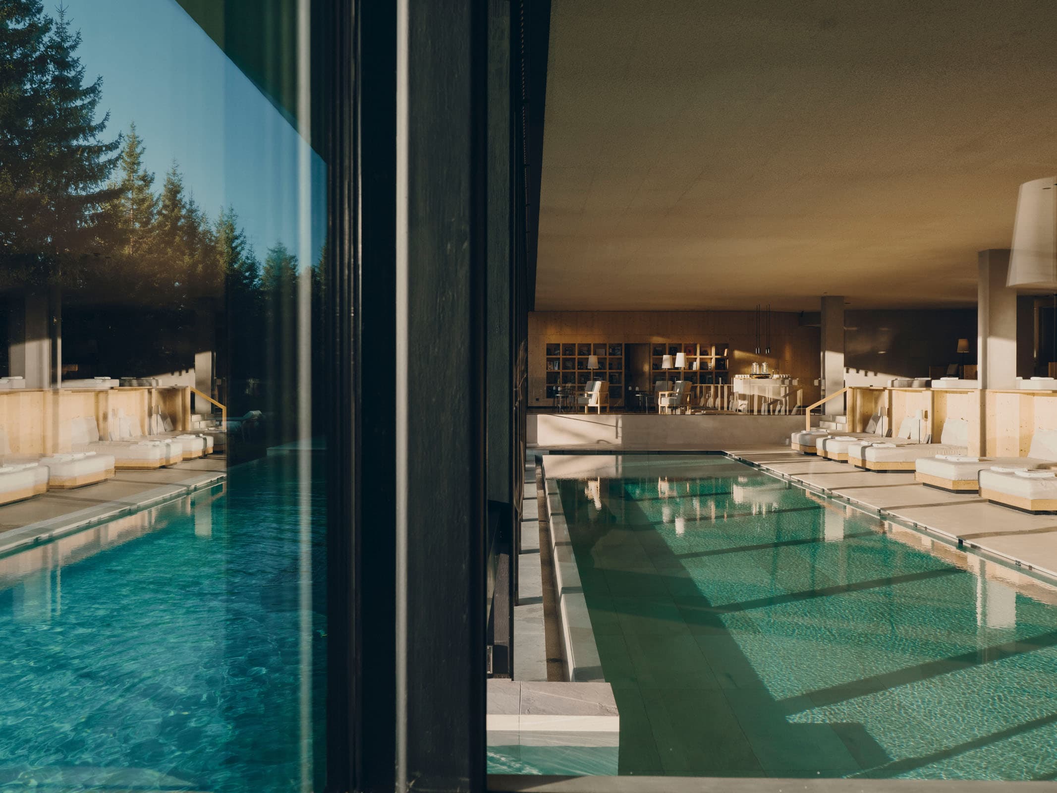 The lap pool seen along its length — pale wooden cabanas down one side, an outdoor pool through the glass at the far end.