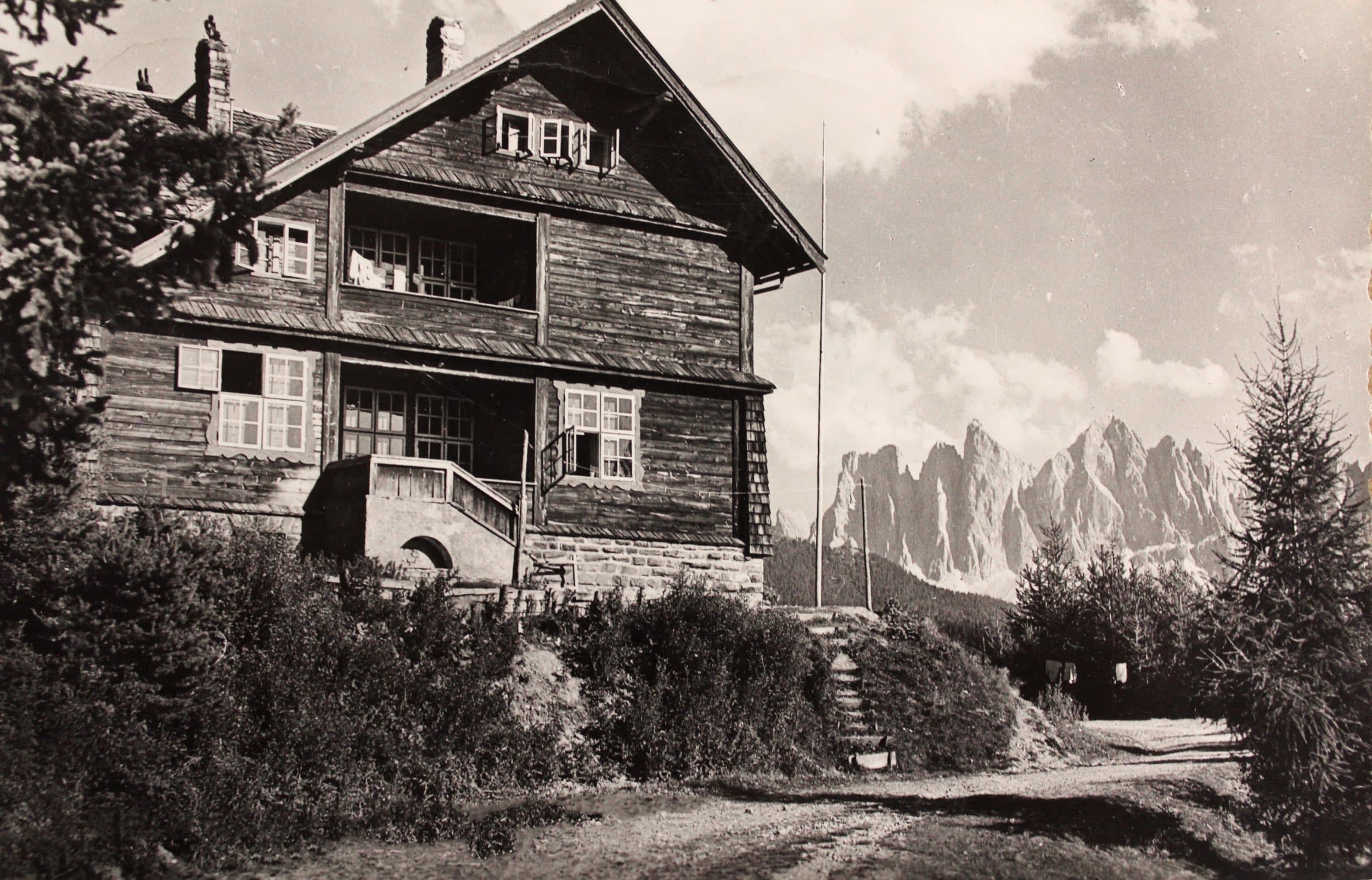 An archival black-and-white photograph of the original 1912 timber sanatorium at Palmschoß — chalet roofs and weathered timber, the Geisler Dolomites rising behind.