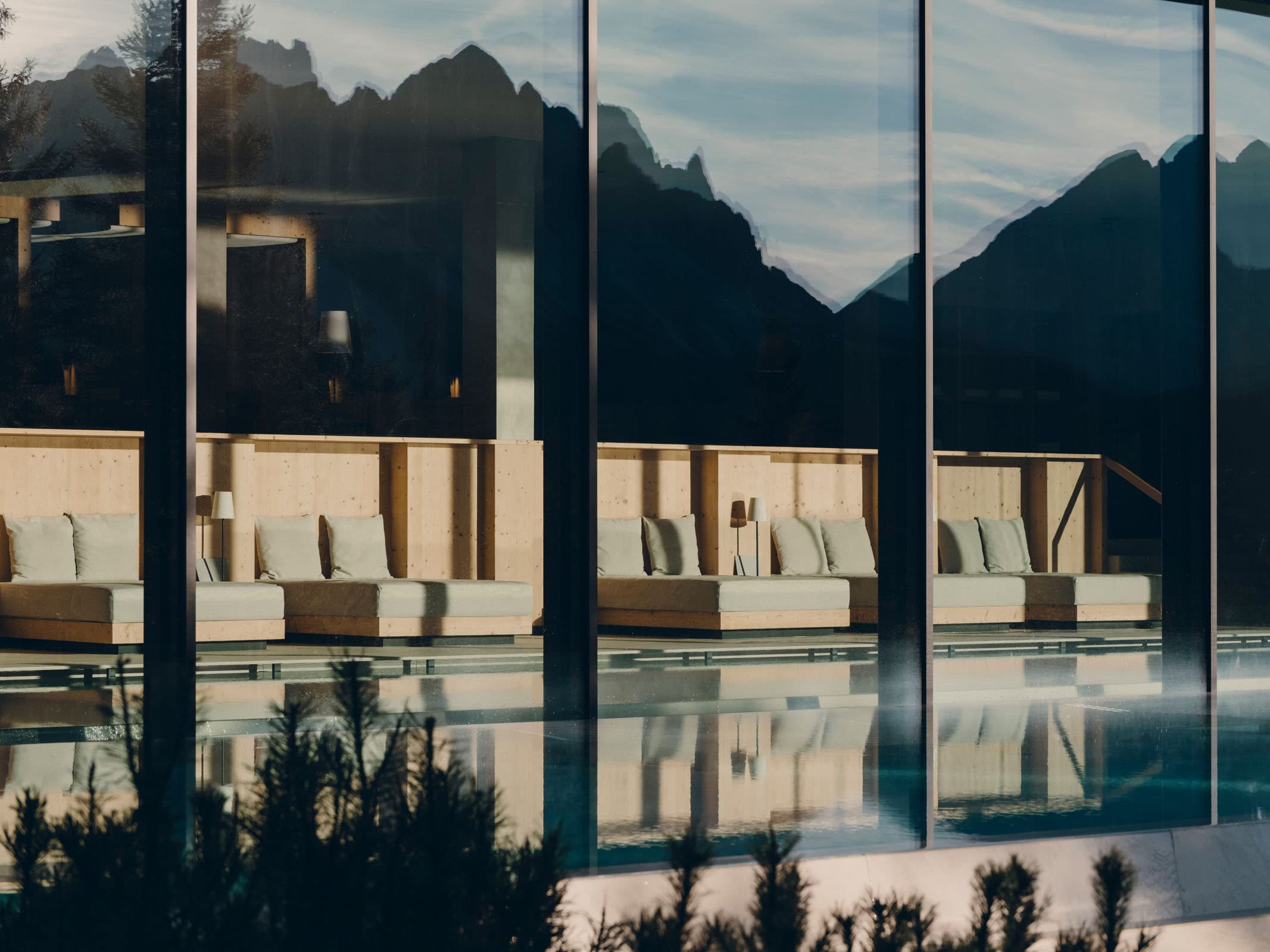 The spa pool seen through a wall of plate glass, the Geisler Dolomites mirrored across its surface, pale wooden loungers on the strip of lawn between water and forest.