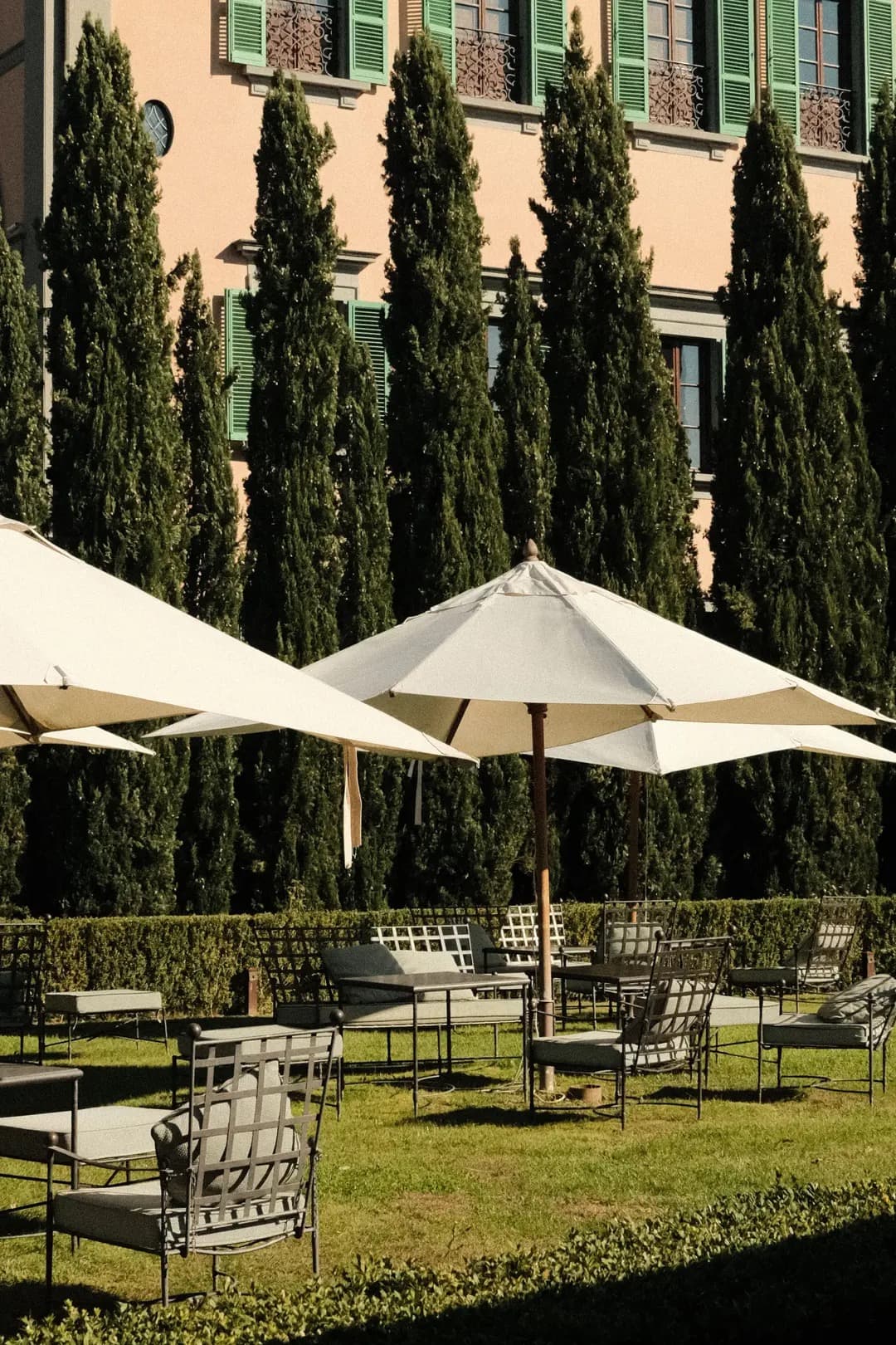 The pink-stuccoed façade of the Dimora Storica with green shutters, framed by tall Italian cypresses and a parterre of cream umbrellas and iron café chairs.