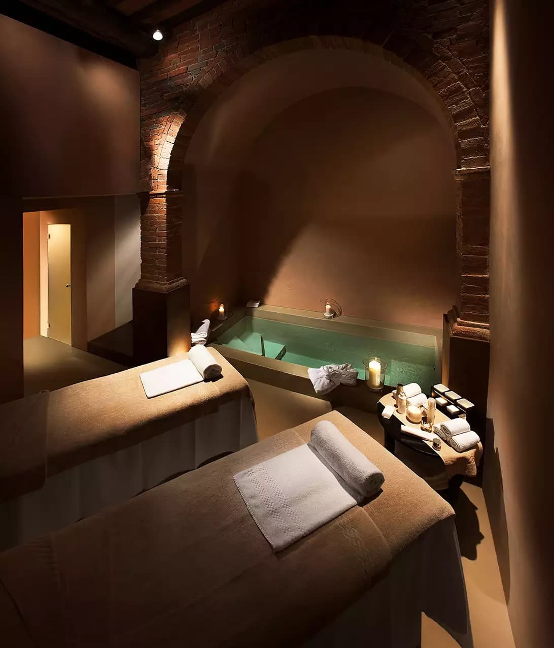 Spa La Corte beneath the village — a small lap pool set under a brick-vaulted arch, two treatment beds laid in candlelight.