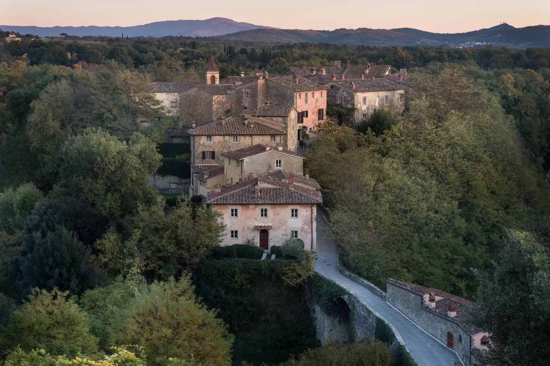 The medieval village of Il Borro at dusk, stone houses clustered on a wooded ridge with the Tuscan hills fading behind.