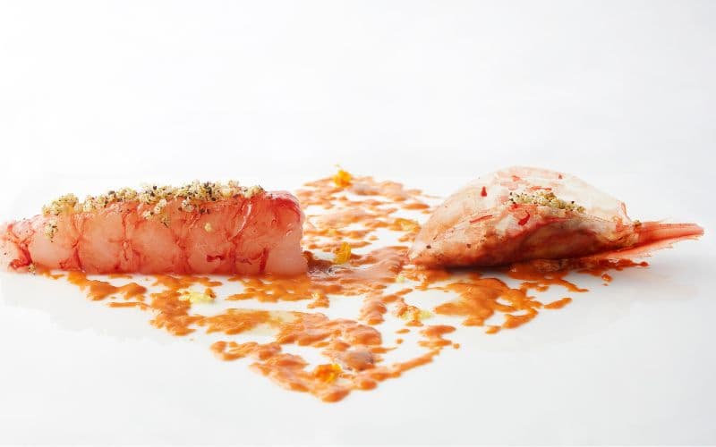 A signature Uliassi plate — a slice of raw Adriatic red shrimp set on a flat white plate beside a pool of orange-flecked sauce and the head of the same prawn, glossy in its own juices.