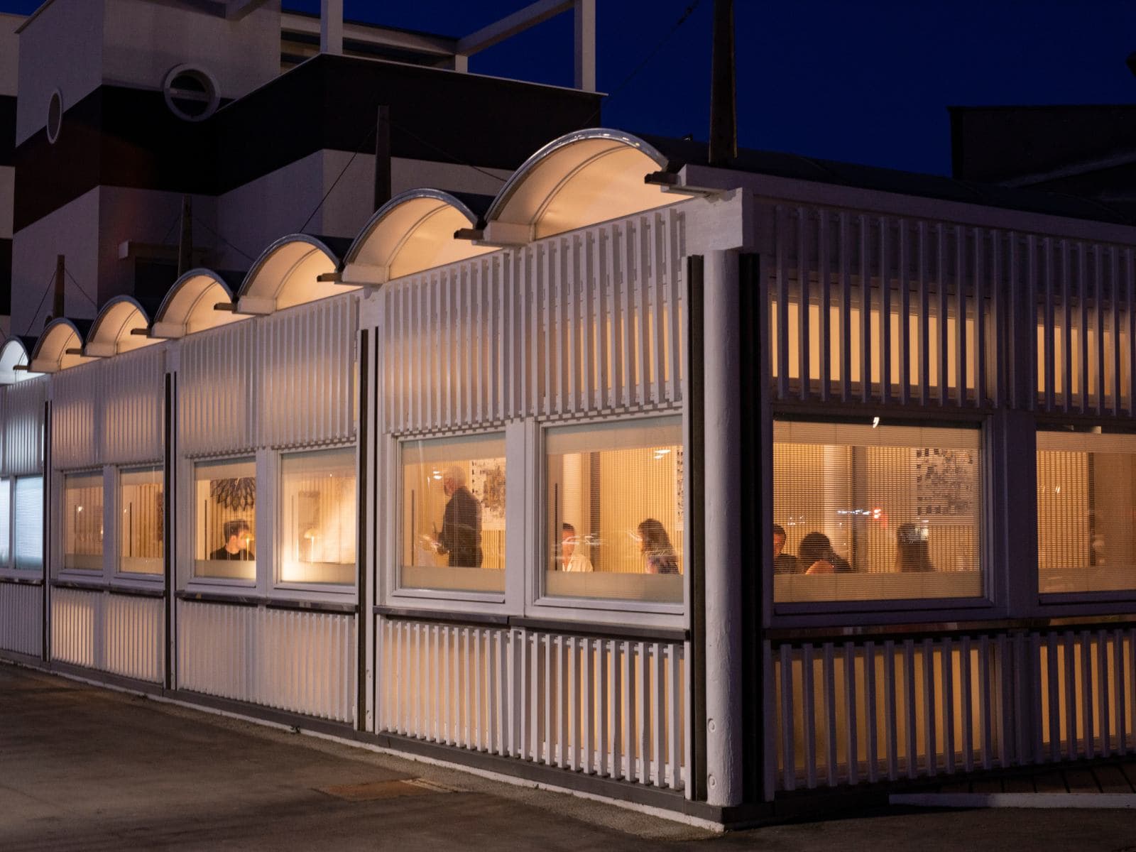 Uliassi at night — the long pavilion lit from within, the arched skylights glowing white, the dining room visible through the windows, a low harbour wall in front and the dark canal mirroring the lights below.