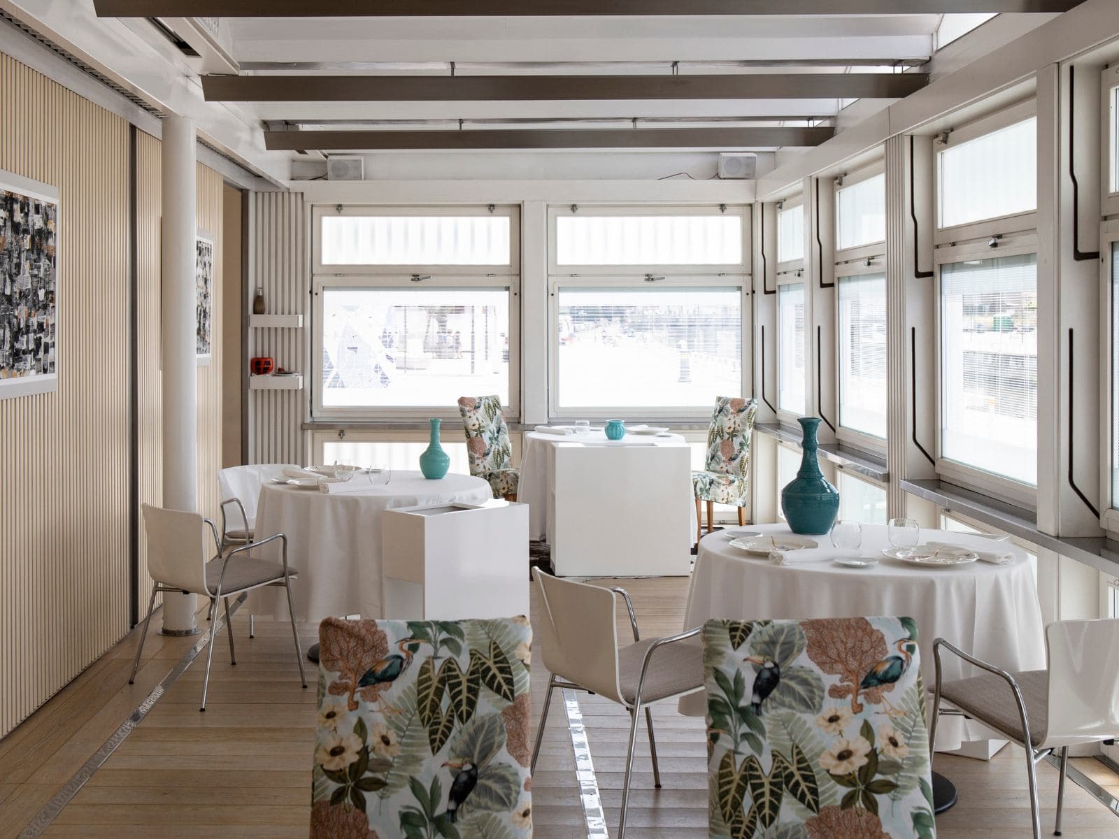 The smaller dining room at Uliassi — three round tables in white linen, floral-printed armchairs, turquoise ceramic vases on the cloths, slatted screens and the harbour glimpsed through the windows beyond.