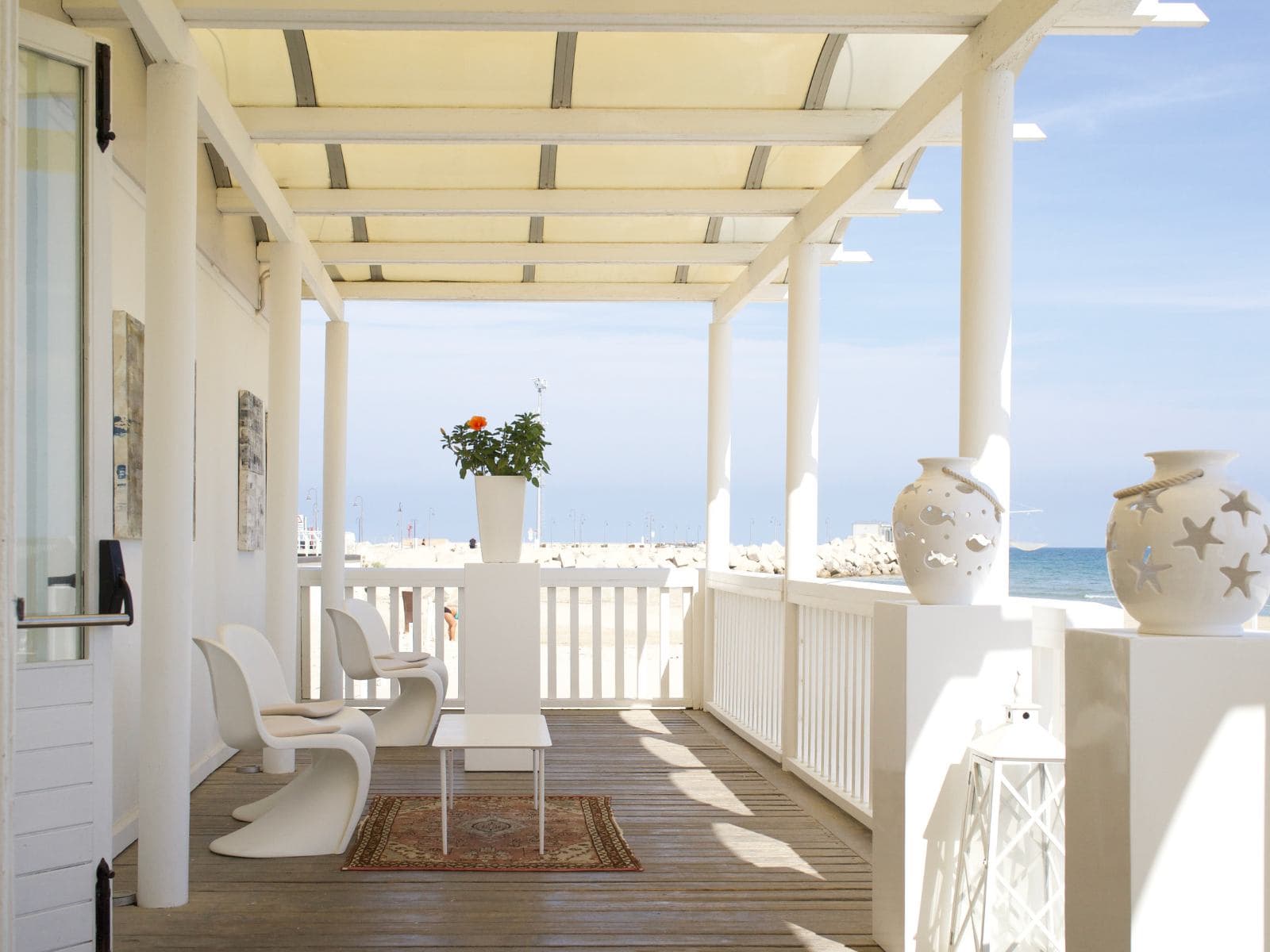 The seafront terrace of Uliassi at midday — white Panton chairs around a low table on a wooden deck, a row of slatted white columns, ceramic urns painted with starfish and fish, the open Adriatic and a strip of sand framed by the white balustrade.