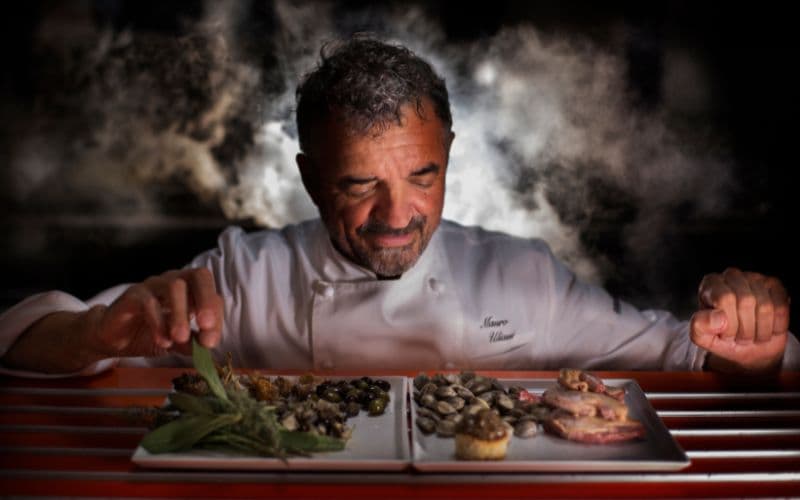 Mauro Uliassi in chef's whites bent low over a long tray of small dishes, steam rising into the dark, his eyes closed in concentration as he places a final garnish.