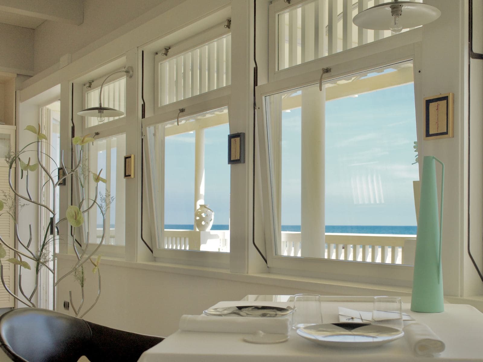 A corner of the Uliassi dining room — a white-clothed table set for two with a turquoise jug and a single black plate, the line of the open Adriatic visible through tall white-framed casement windows along the sea-side wall.
