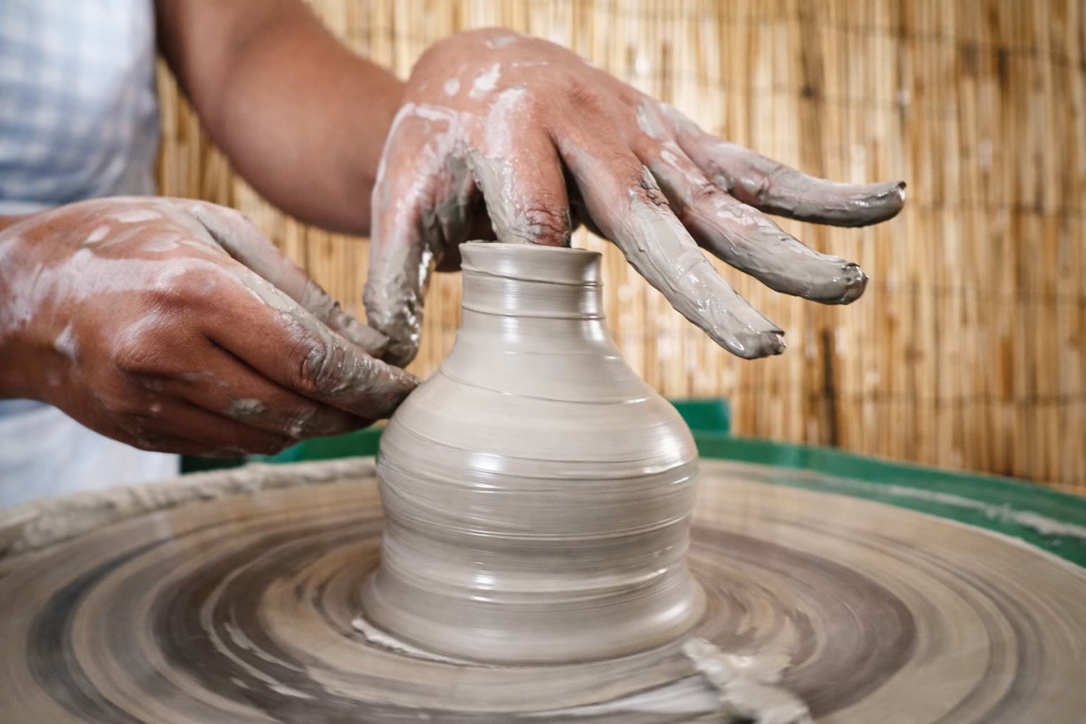 Close view of two clay-covered hands shaping a tall vessel on a spinning potter's wheel, reed screen behind.