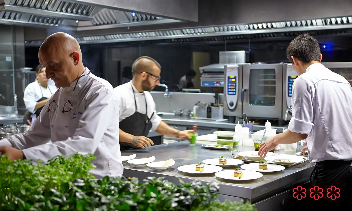 The pass at Atrio — three chefs in whites finishing a row of plates, a low rampart of fresh herbs in the foreground, the three Michelin stars marked at the corner of the frame.