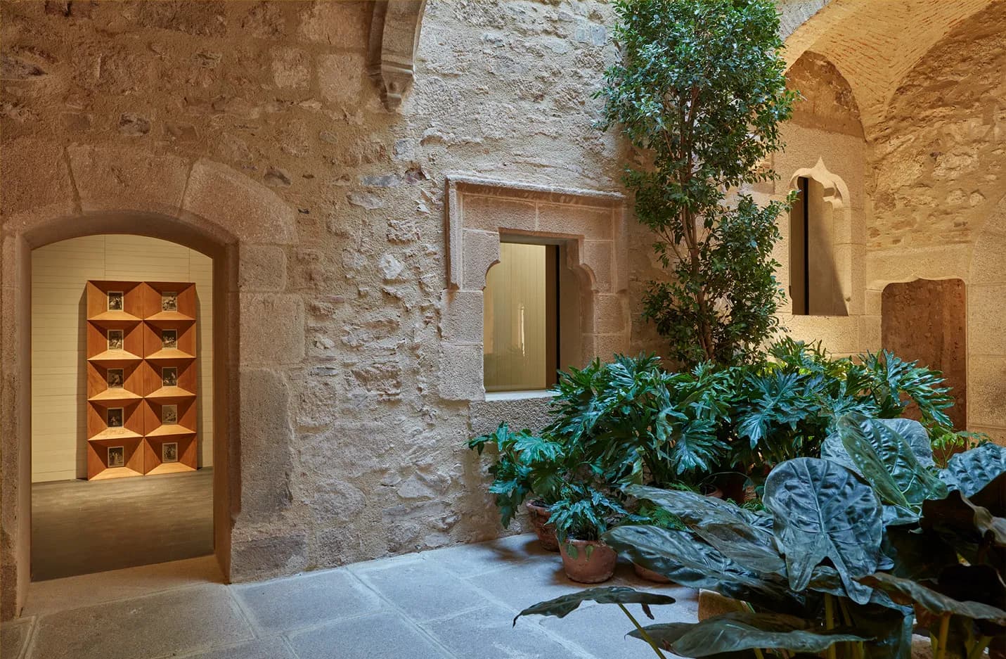 A small inner courtyard of the palace — pale granite walls under a vaulted arcade, a single tree and a row of philodendron in terracotta, a recessed bronze niche on the back wall.