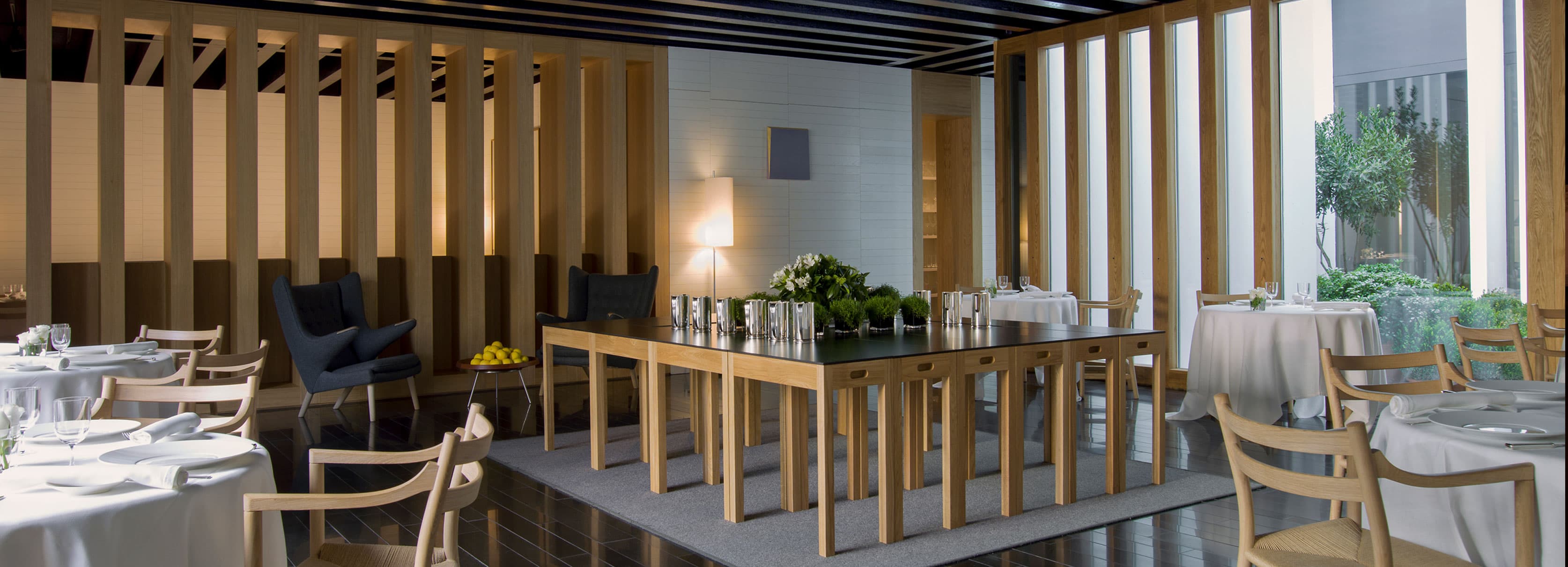 The Atrio dining room — a long room of vertical timber fins, white-clothed tables and pale chairs, opening through full-height glass to a stone-walled garden.