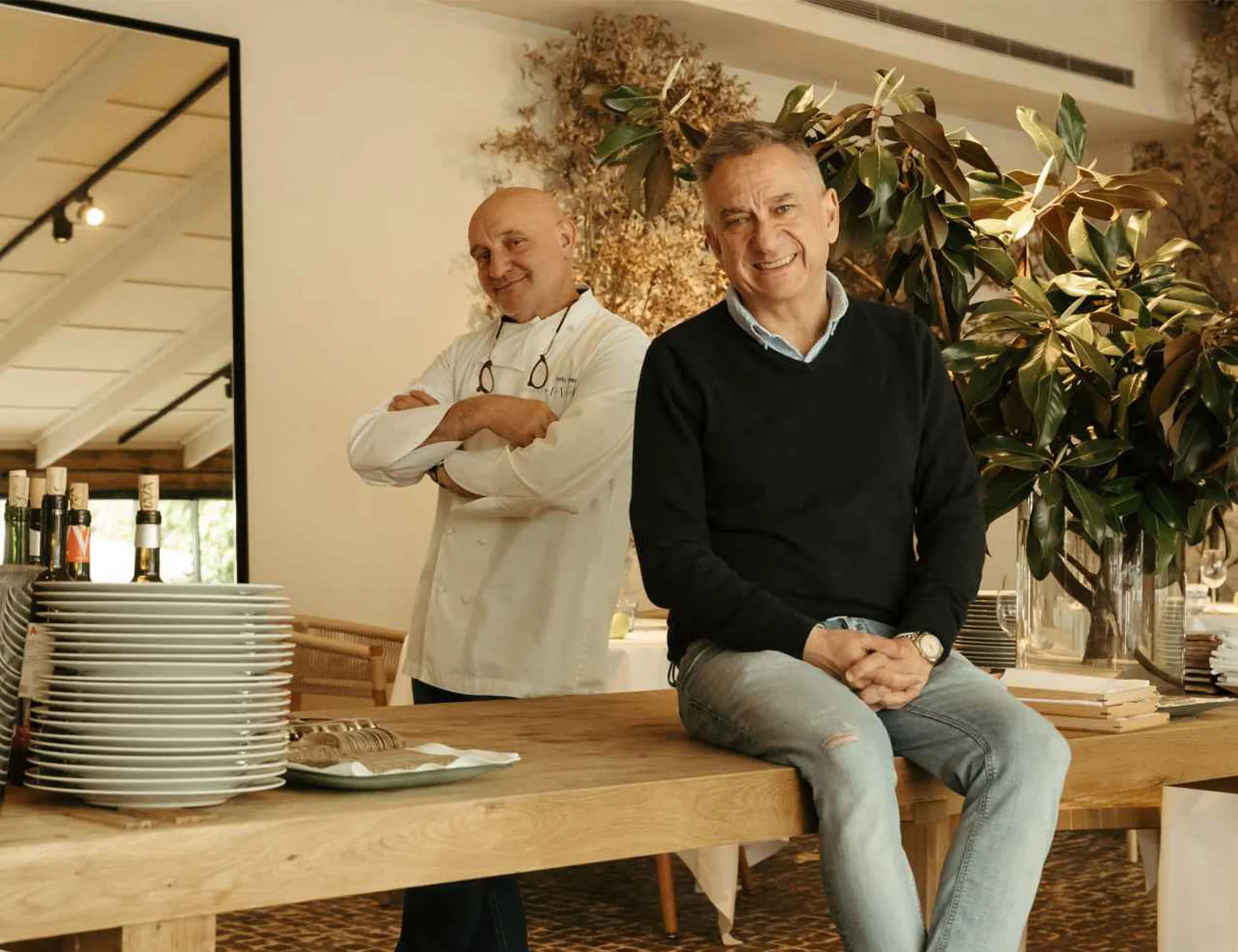 Toño Pérez in chef's whites and Jose Polo in a dark sweater, photographed beside a long timber table piled with white plates and a bowl of magnolia leaves.