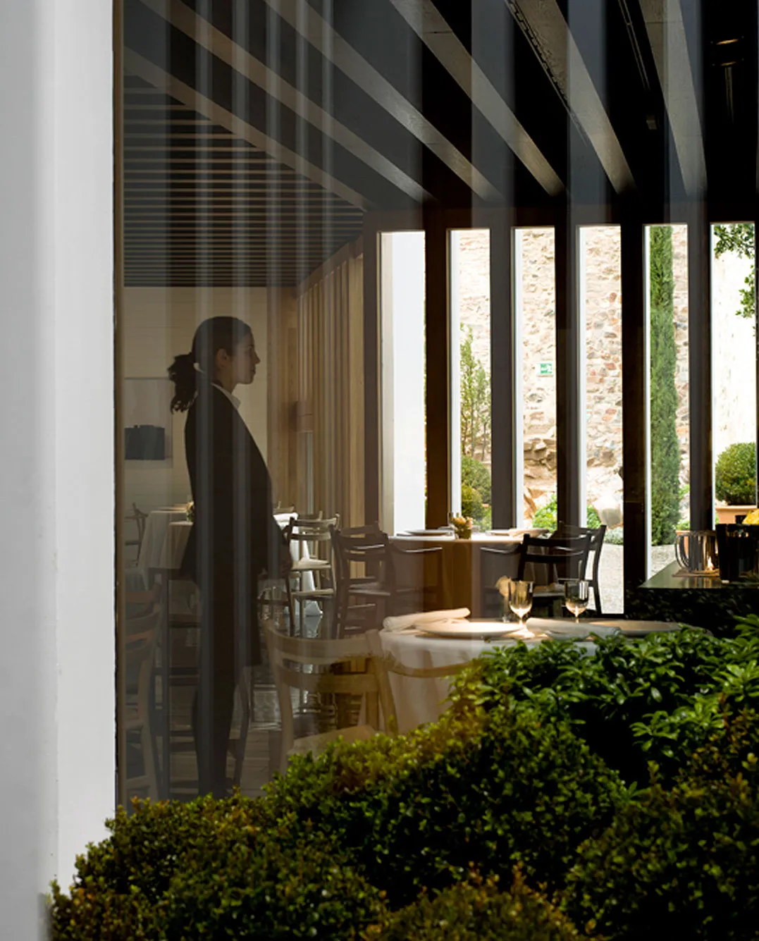 A view through the dining-room glass — a sommelier in silhouette in front of vertical timber fins, white-clothed tables set for service, a stone-walled garden with cypress and topiary beyond.