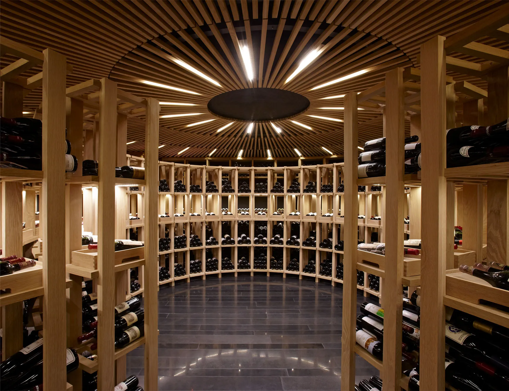 The circular wine cellar — pale timber columns radiating from a central oculus, deep shelves stacked floor to ceiling with bottles.