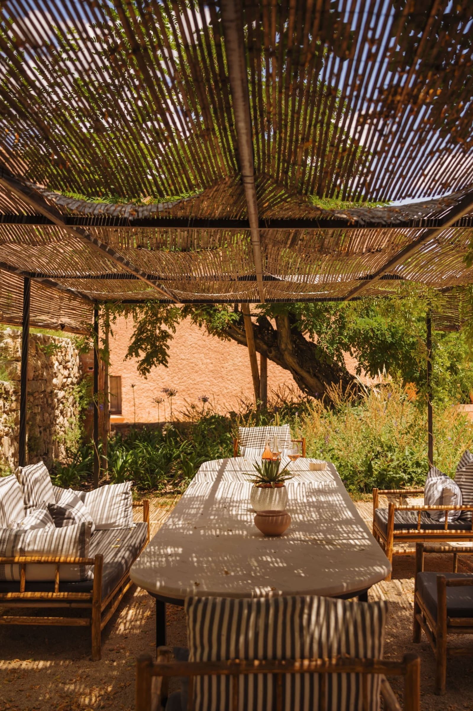 The outdoor pergola at midday — a long trestle table under a woven cane shade, striped banquettes along one side, the courtyard's stone wall and a wide tree behind.