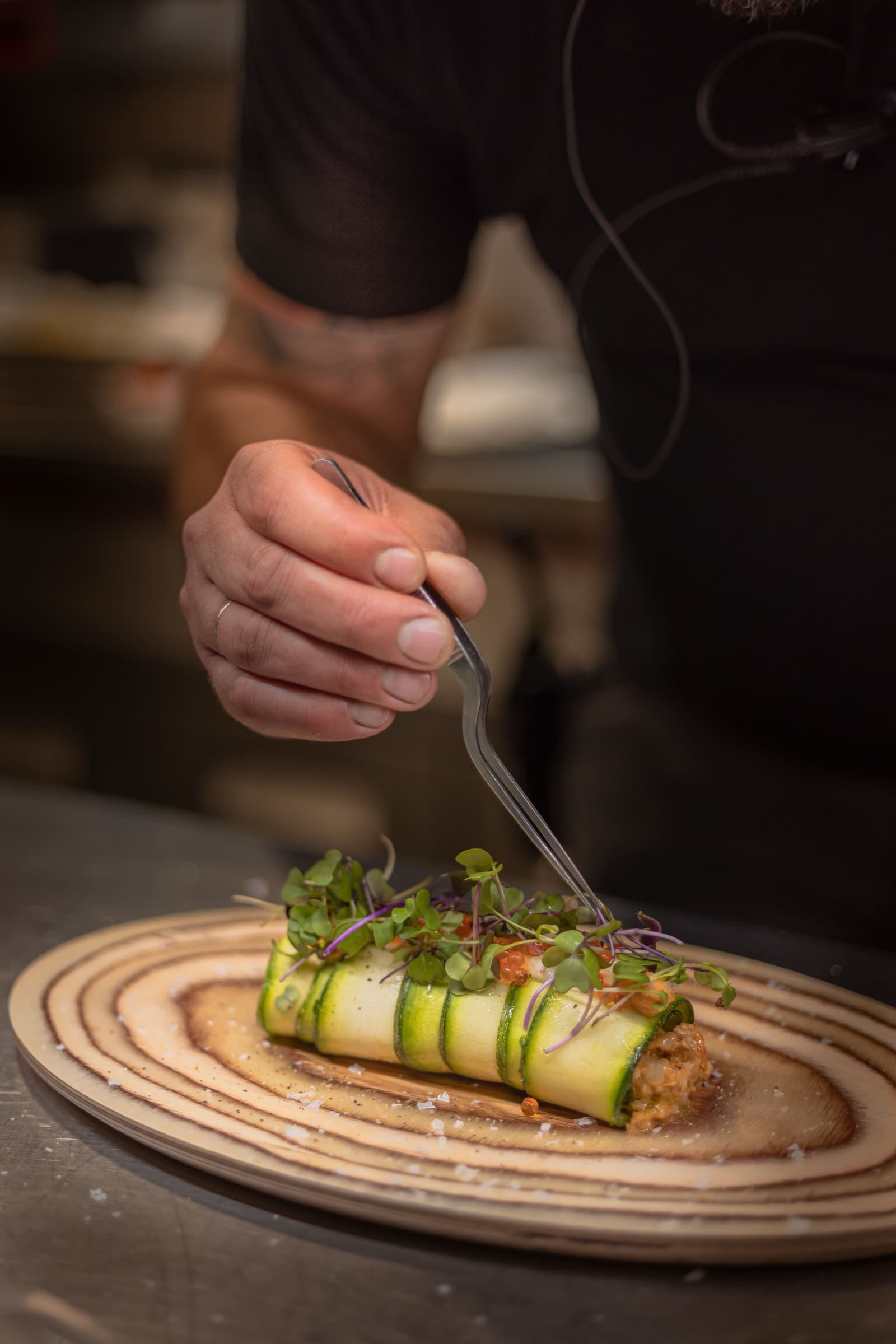A close shot of a courgette-ribbon roll being plated on a sliced wooden disc — the chef's hand placing micro-herbs on top with tweezers.