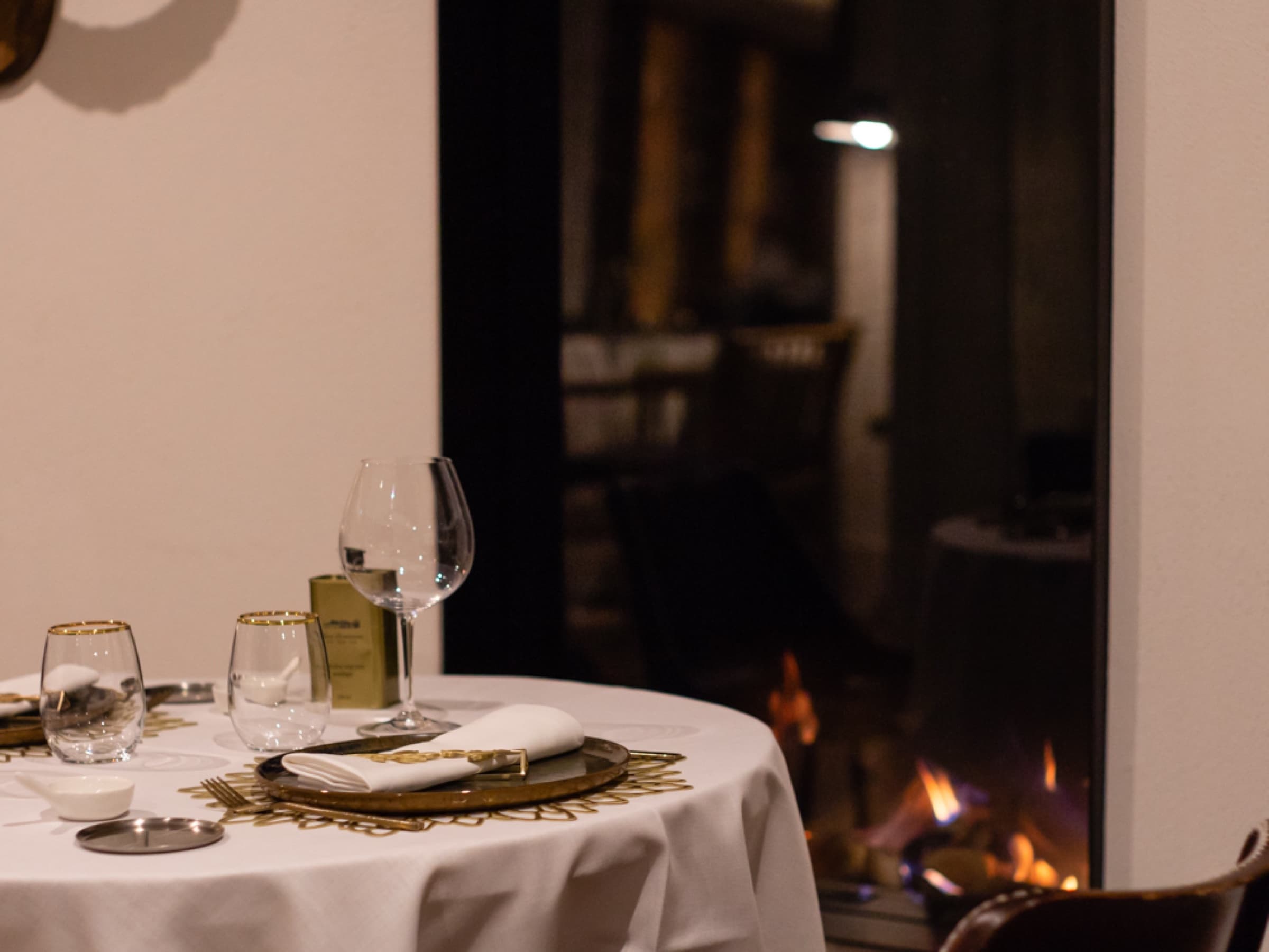 A round table dressed in white linen with gilt-rimmed glassware and a sun-disc place setting, an open fire glowing in the hearth across the room.