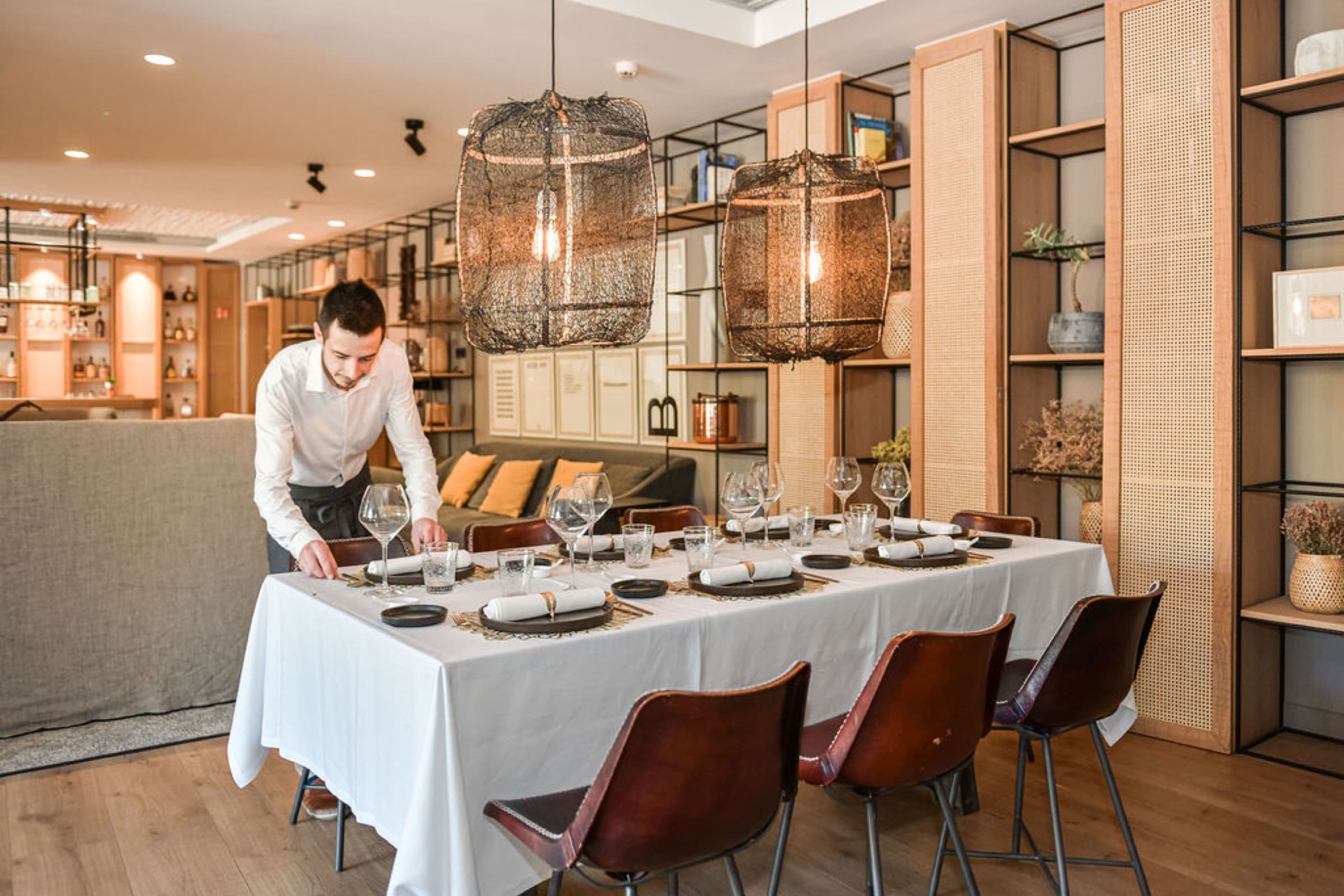 A waiter in a white shirt setting a long table for the tasting menu, two large woven black pendants overhead, caned shelving along the wall, leather chairs on the warm wood floor.