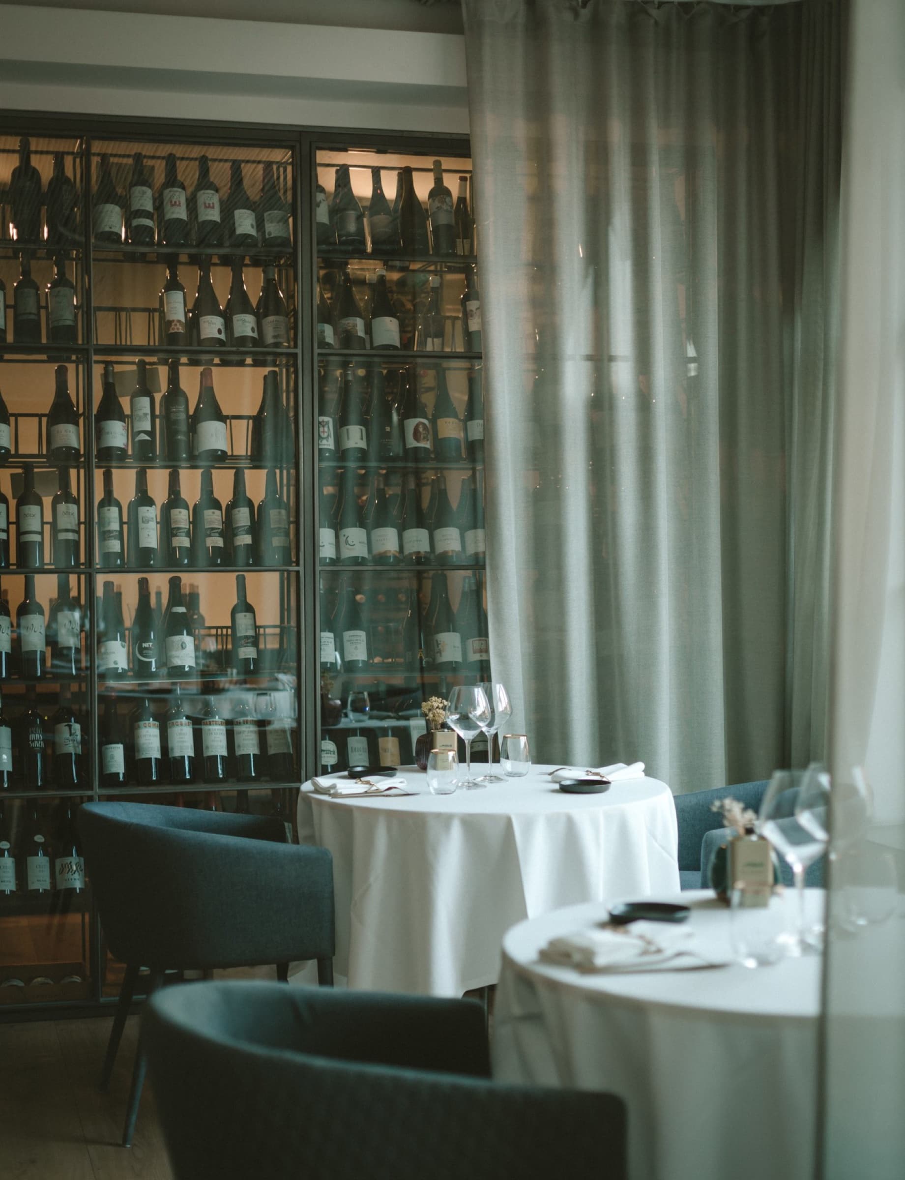The Mater Terrae dining room behind a floor-to-ceiling glass wine cellar — backlit bottles in dark steel racks, a sheer linen curtain, white-clothed round tables and slate-blue armchairs in front.