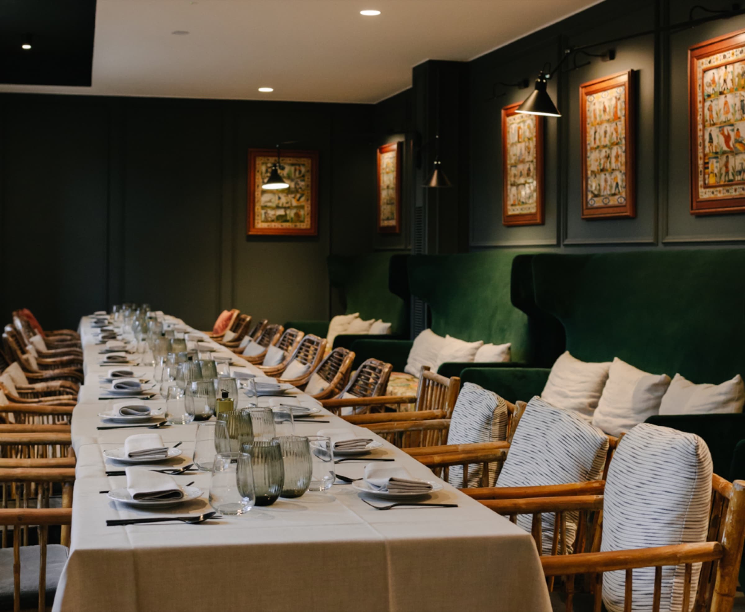 A long communal table in a deep racing-green dining room — green velvet banquette, framed cartographic prints under warm wall lamps, bamboo armchairs along the table set with linen and stemware.
