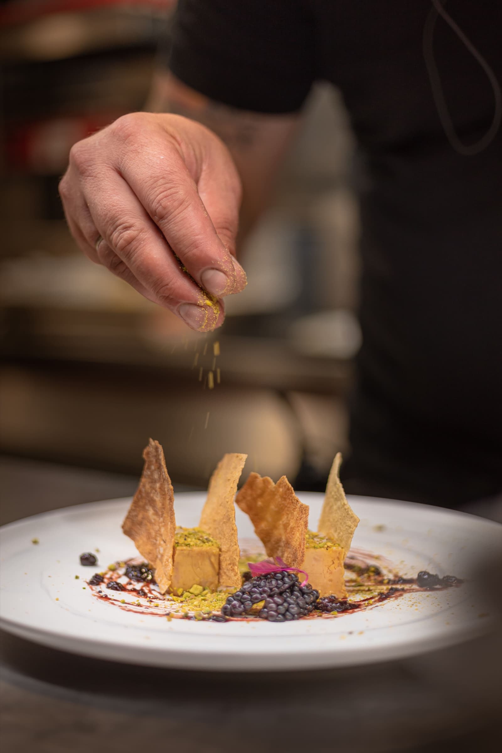 A close shot of the chef's hand sprinkling pale pollen over an arrangement of crisp golden tuiles and dark fruit on a white plate.